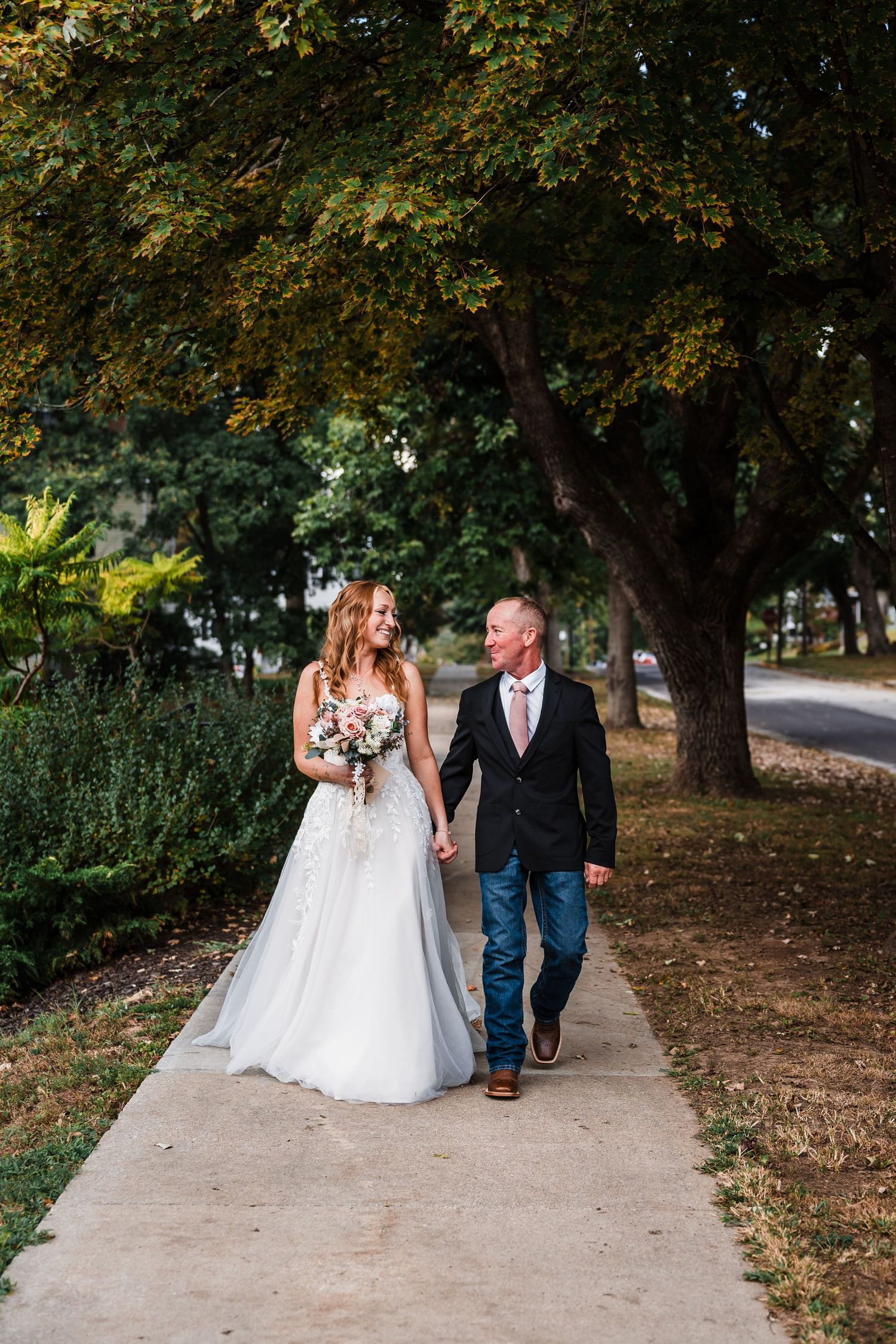 Couple walking together on a tree-lined pathway in wedding attire.