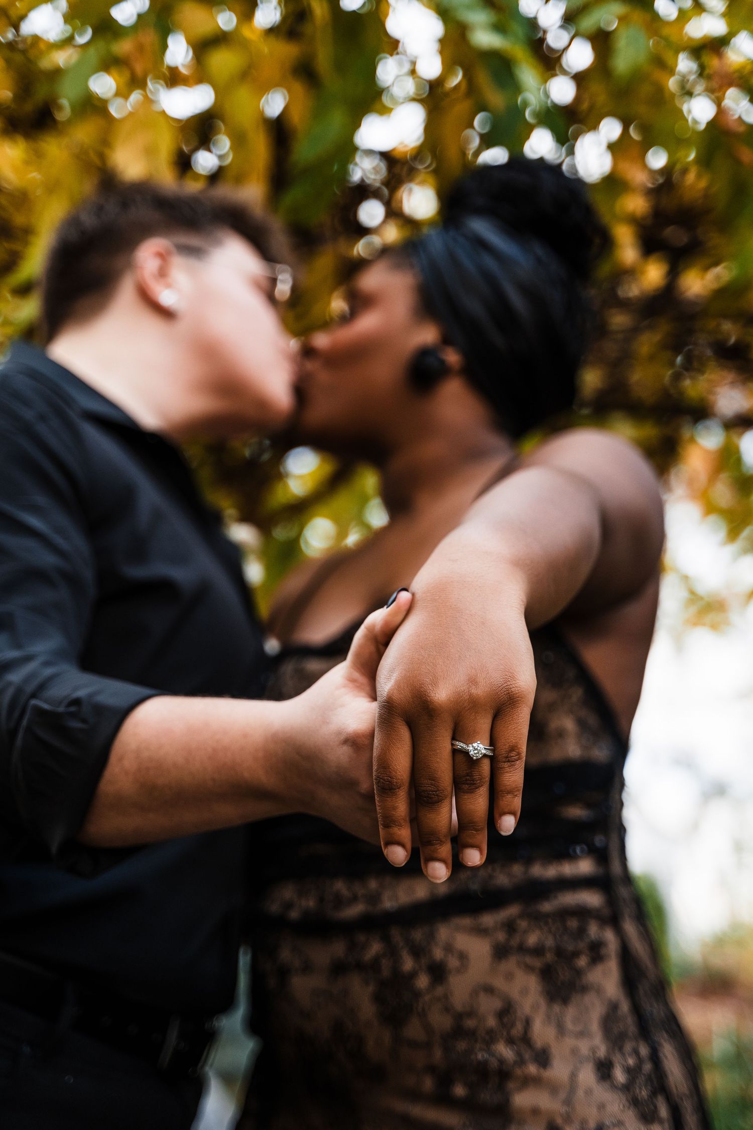 Close-up of two hands with wedding rings as couple shares an intimate kiss under autumn trees.
