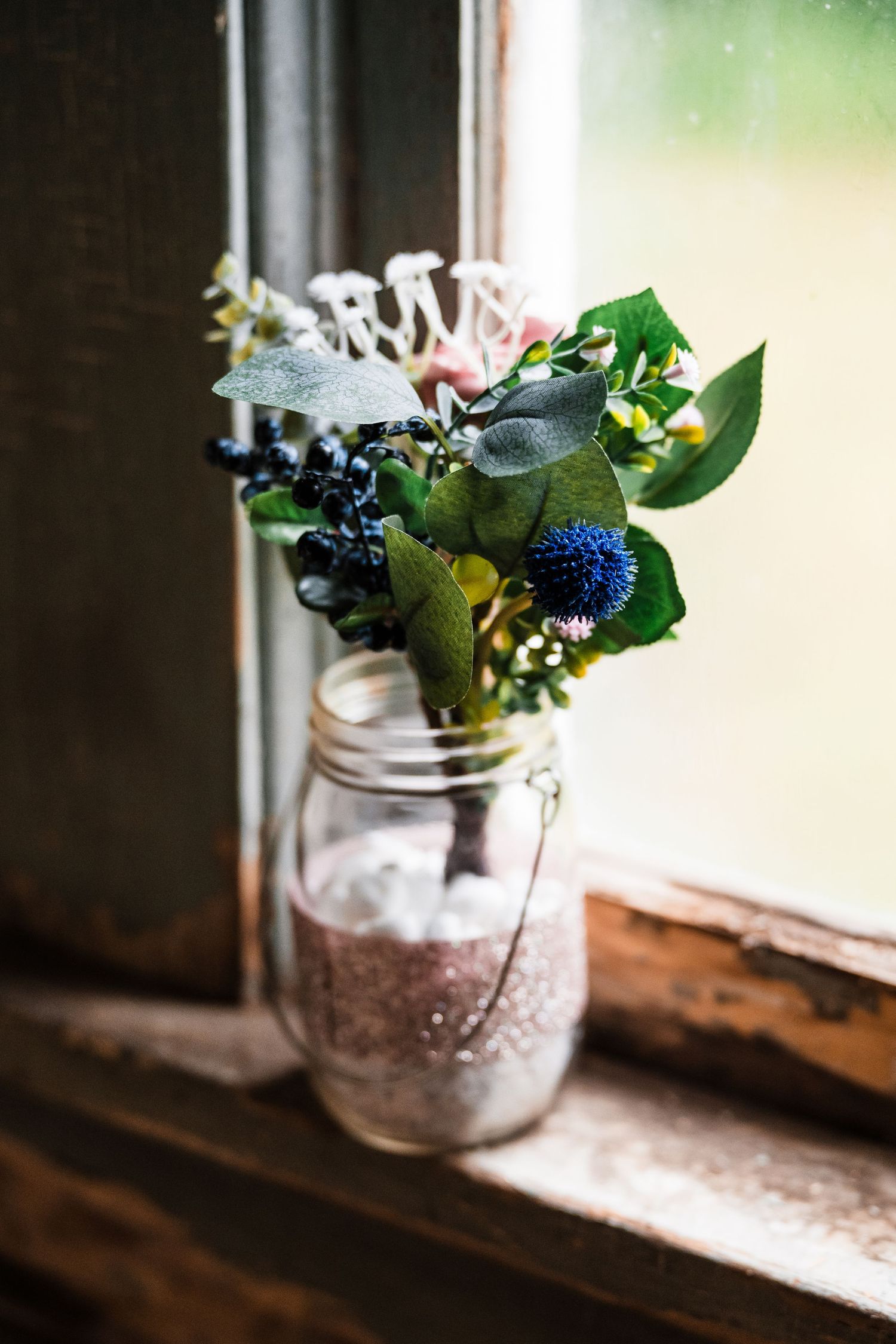 Small glass jar with delicate blue and white flowers sits on rustic windowsill.