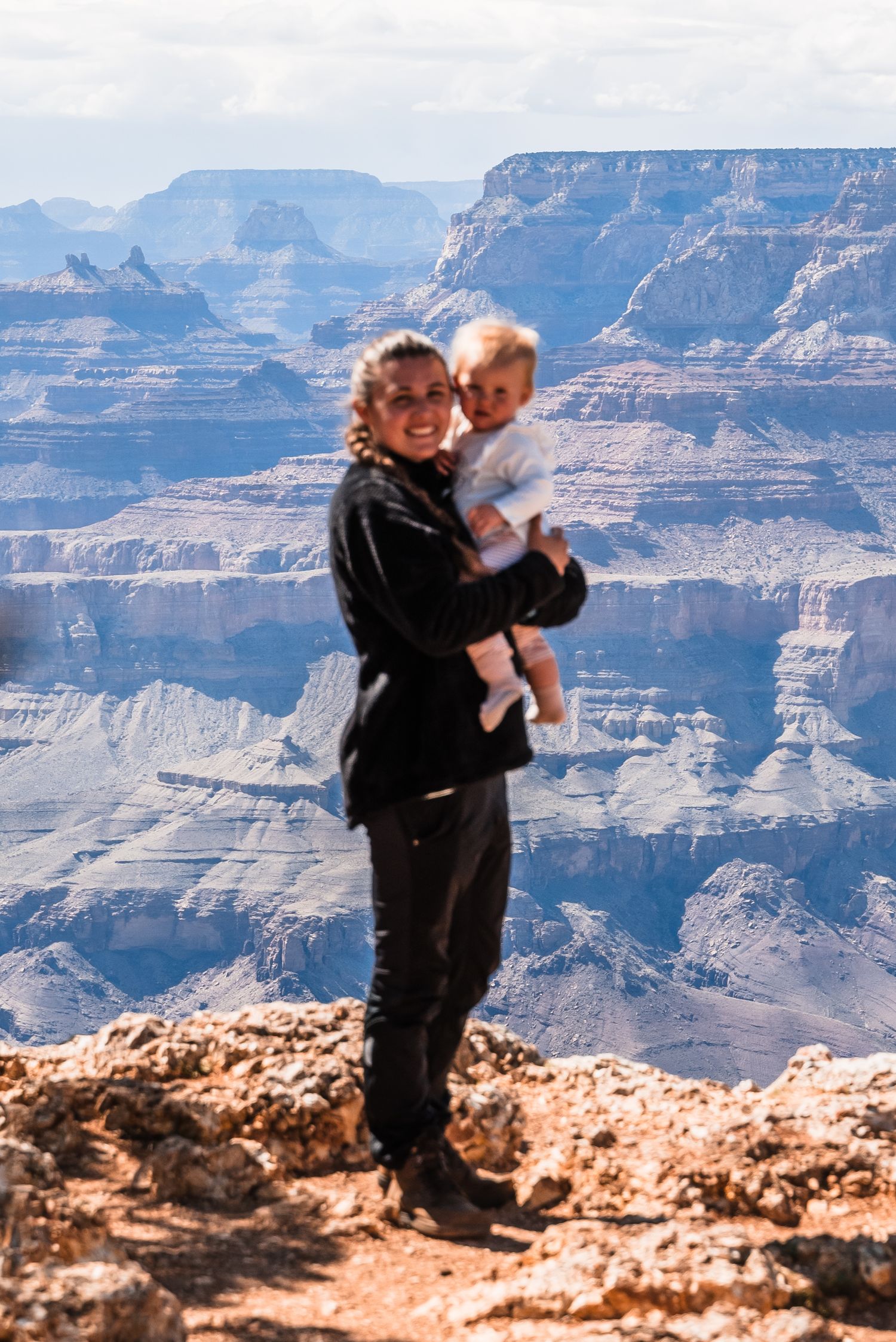 A parent and child stand at a scenic Grand Canyon overlook against dramatic layered cliffs and desert landscape.