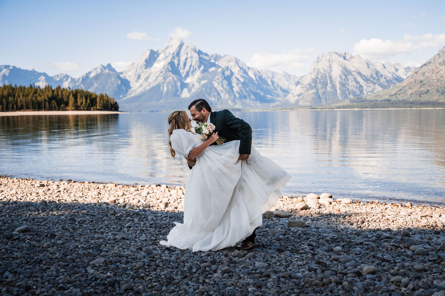 A romantic wedding photo on a rocky beach with snow-capped mountains and a pristine lake in Grand Teton National Park.