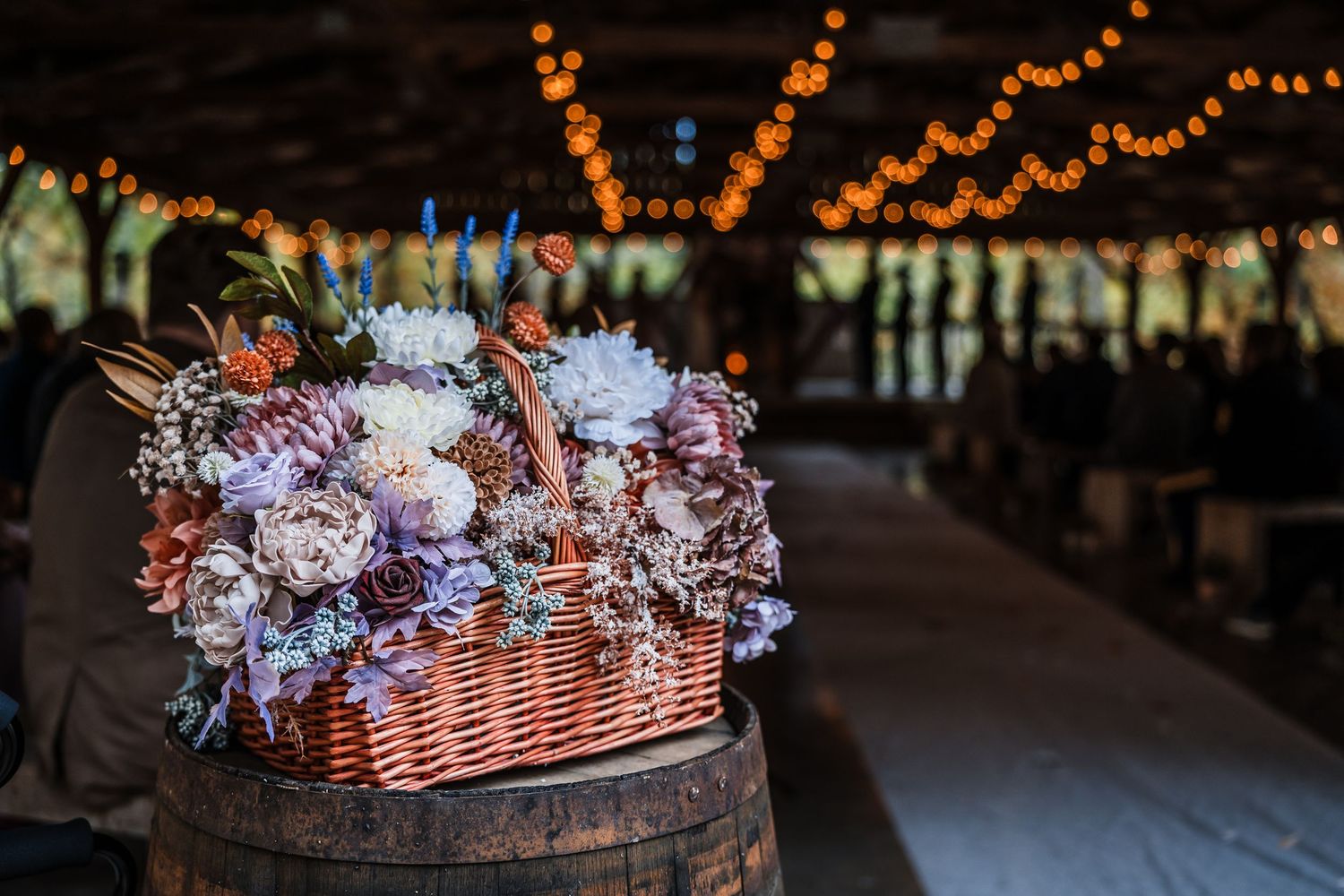 Rustic basket filled with purple and pink flowers on a wooden barrel under string lights.