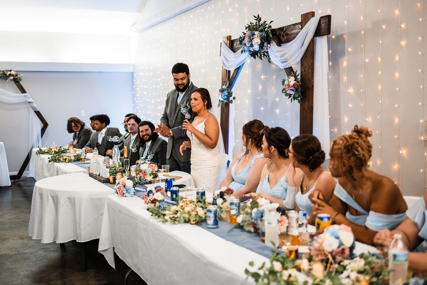 Wedding reception head table decorated with flowers and string lights as guests give toasts.