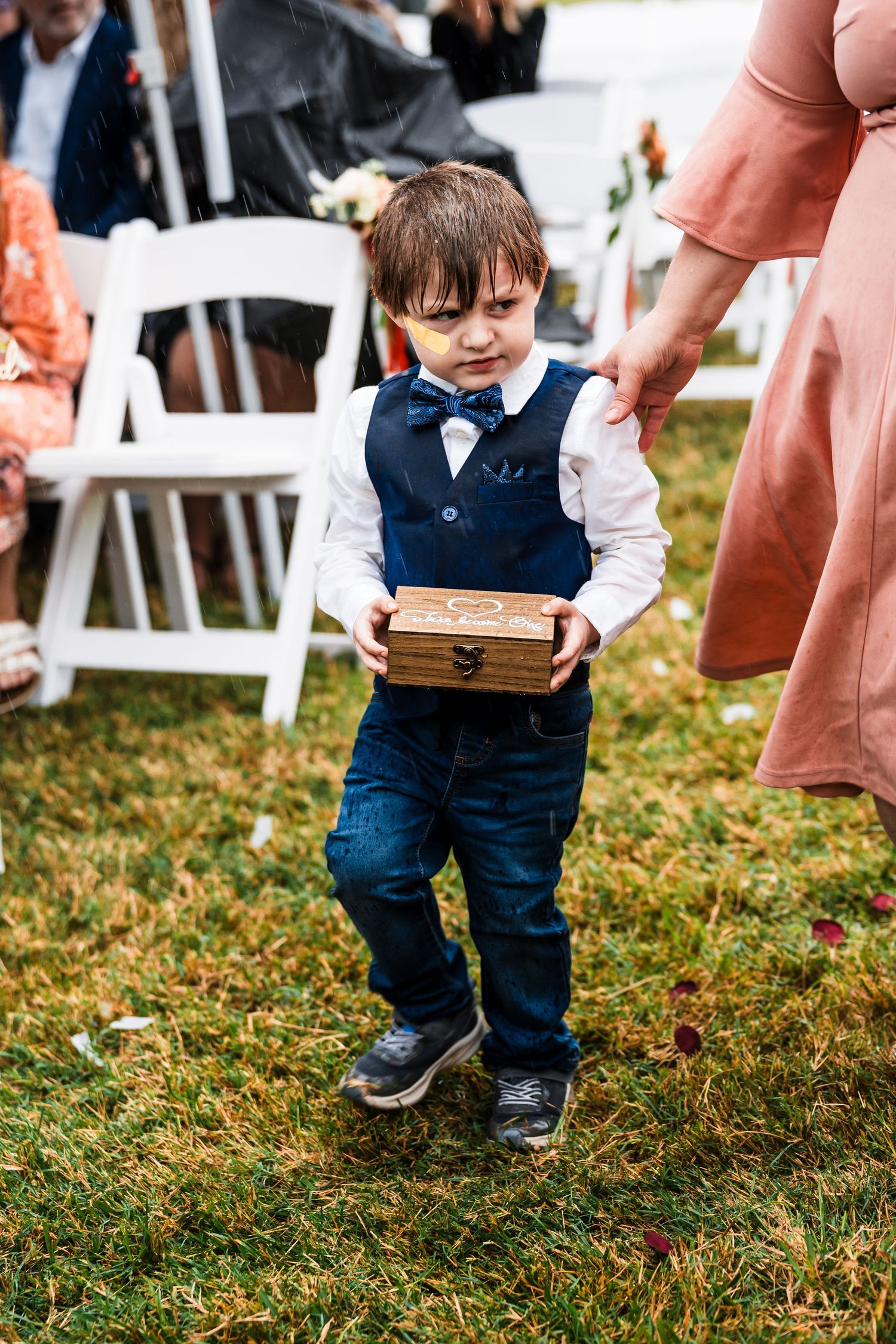 Small child in formal wear walking on grass at outdoor wedding with folding chairs visible.