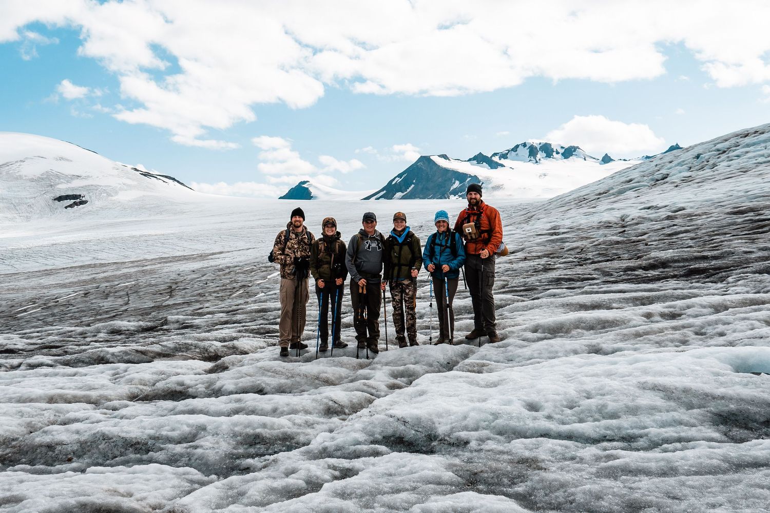 A group of hikers stand together on a snowy glacier field with mountain peaks and blue sky in the background.