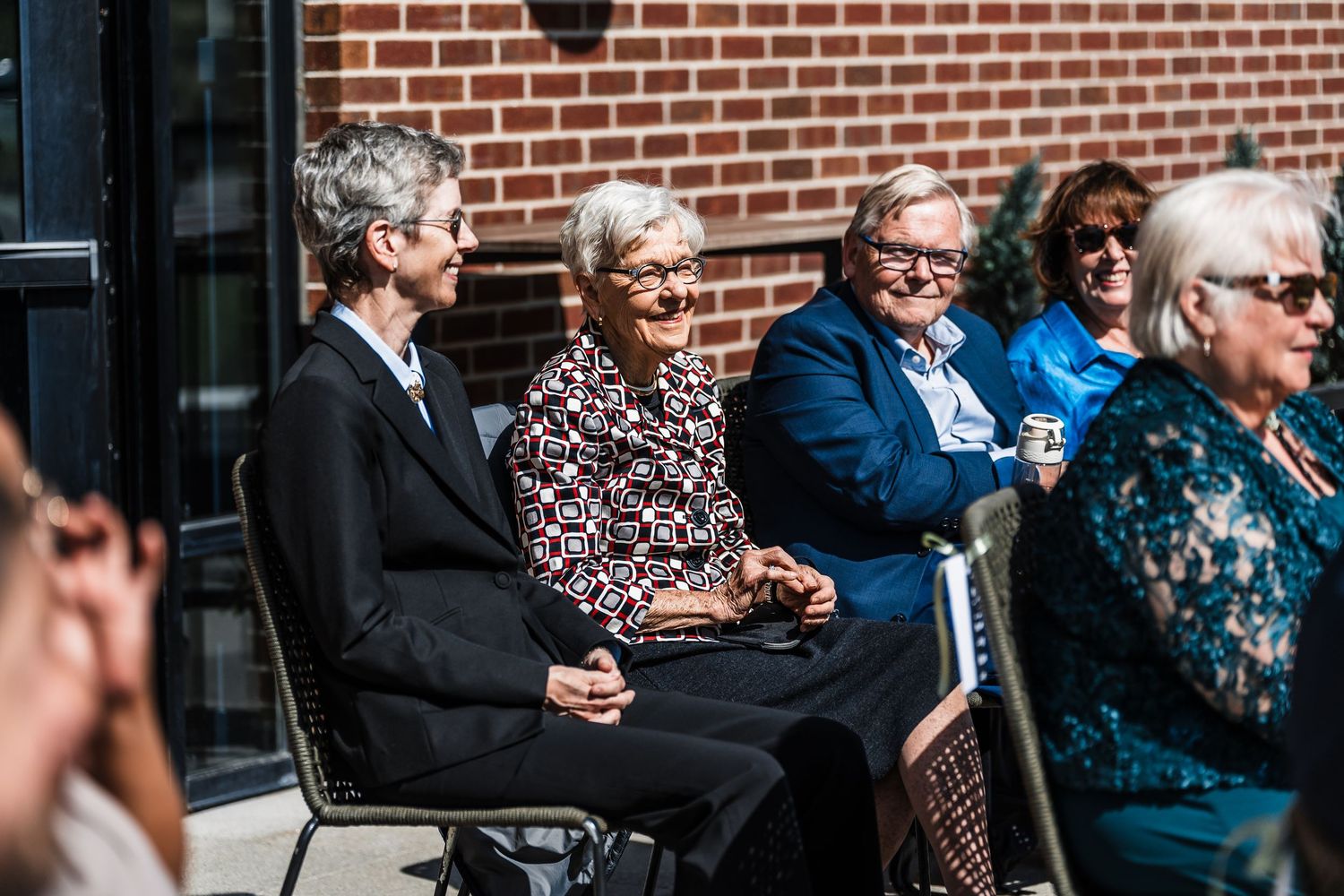 Elderly guests smile and laugh while seated outdoors at a sunny wedding ceremony.