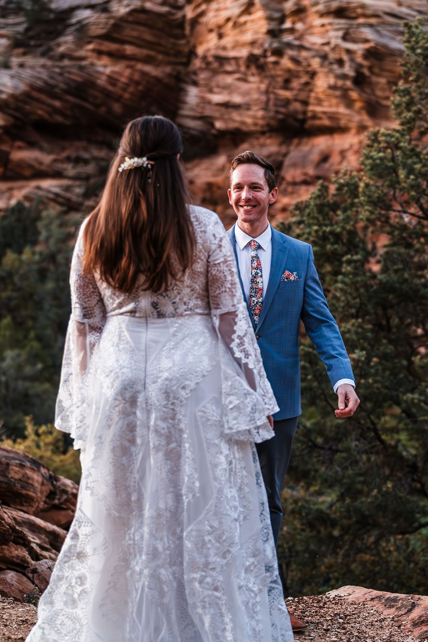 Bride in lace gown and groom in blue suit share a romantic moment among red rock formations.