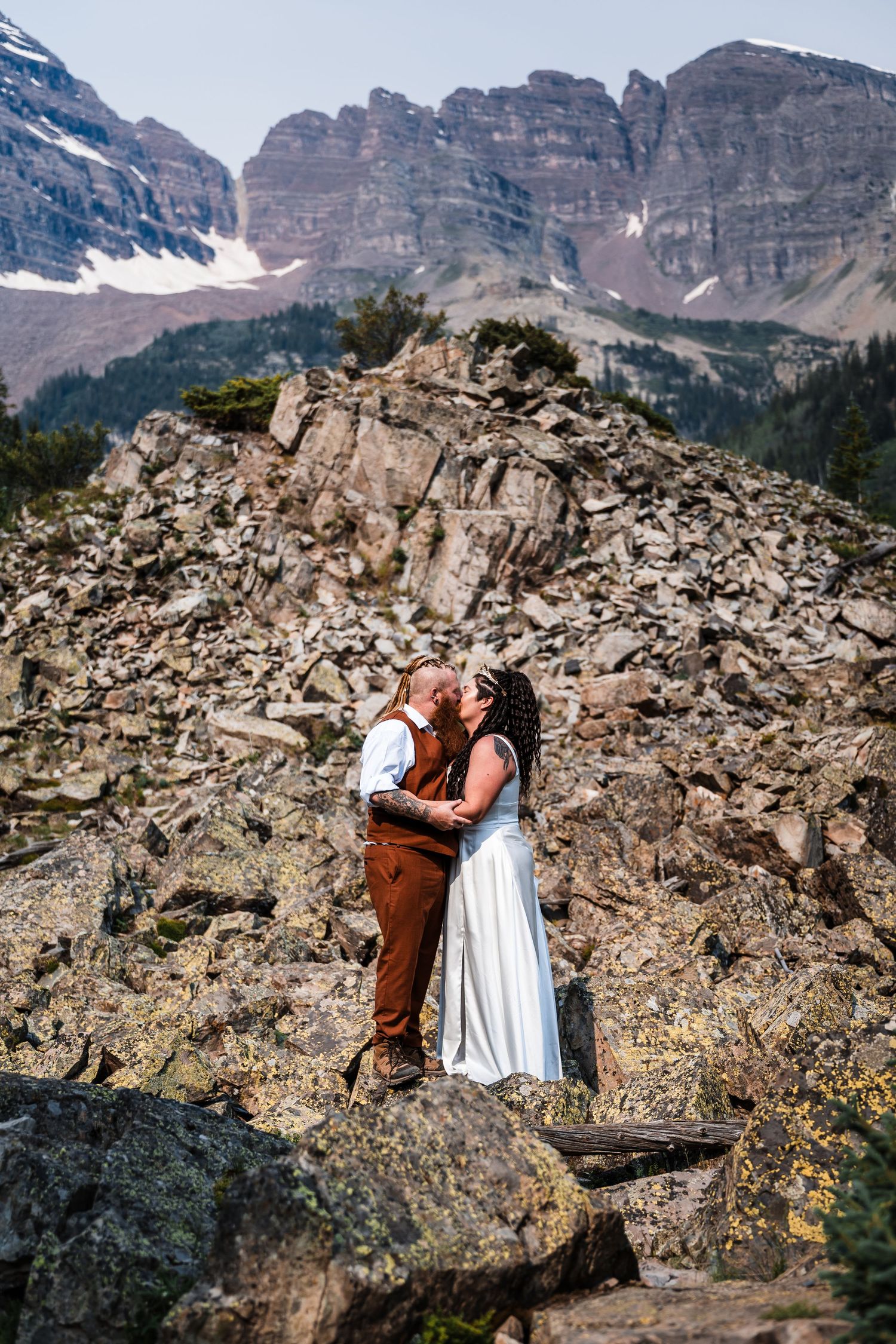 Tattooed bride and bearded groom share kiss on rocky mountain terrain with Maroon Bells peaks in background.