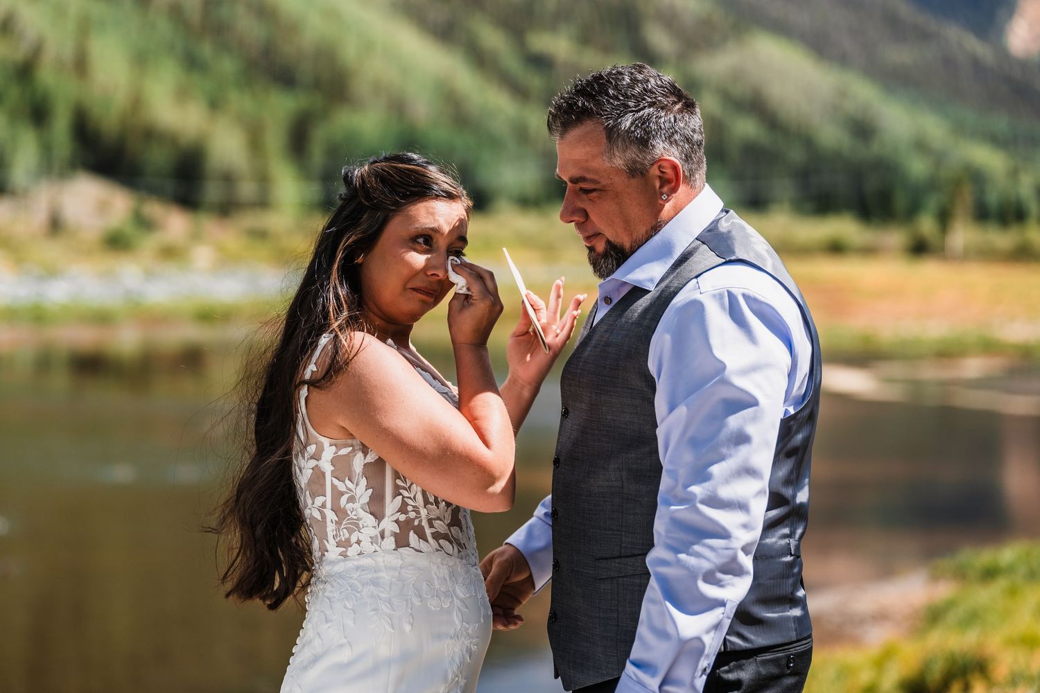 Bride tenderly wipes groom's tears during an emotional outdoor wedding ceremony beside a mountain lake.