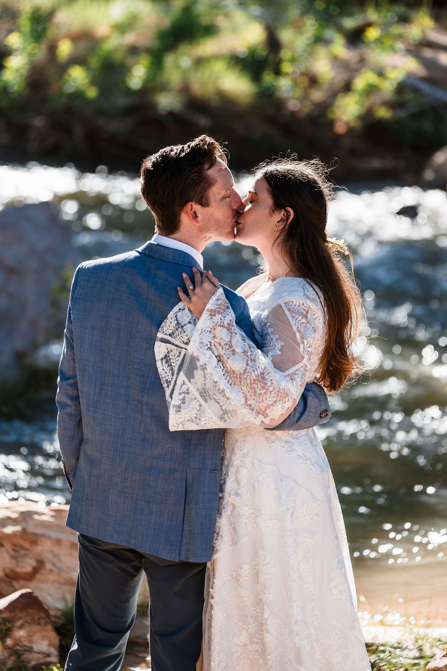 Bride and groom sharing a kiss beside a sparkling river, bride in lace gown and groom in blue suit.
