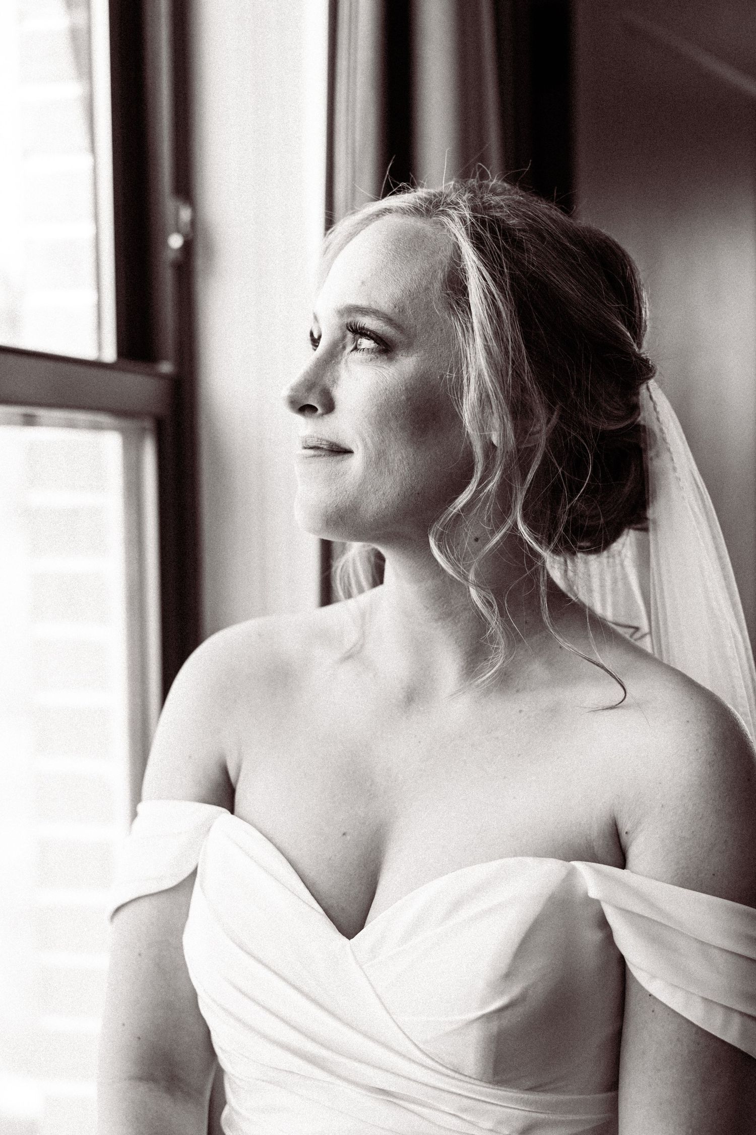 Black and white portrait of bride in off-shoulder gown and veil gazing out a window.
