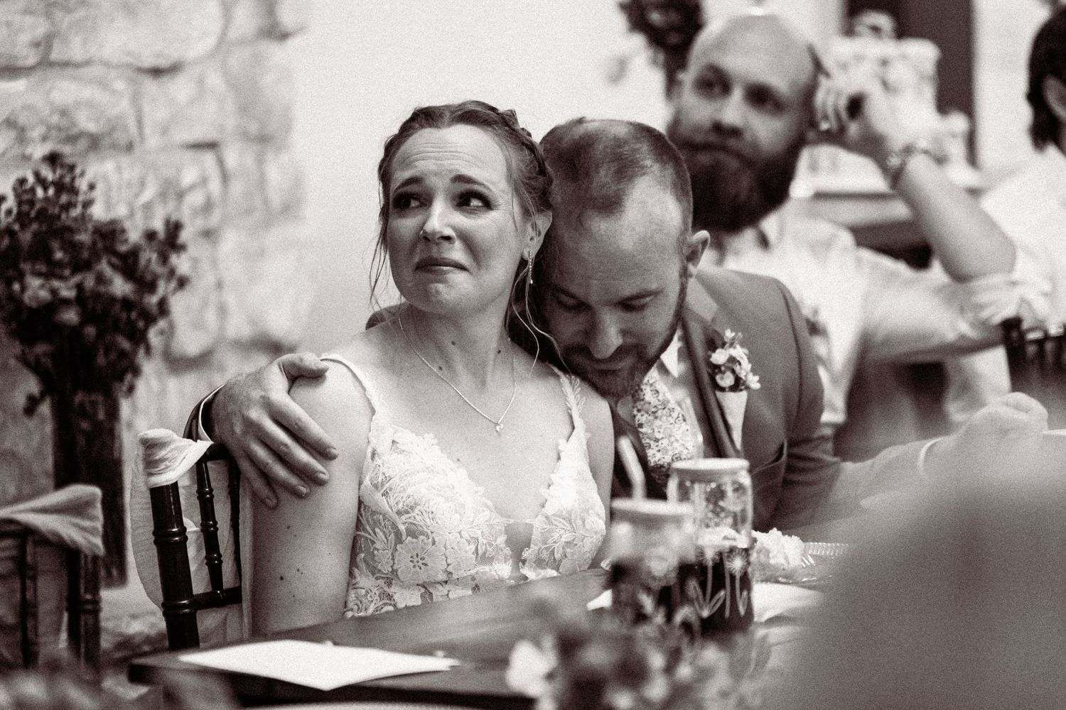Black and white photo of emotional bride and groom embracing at wedding reception table.