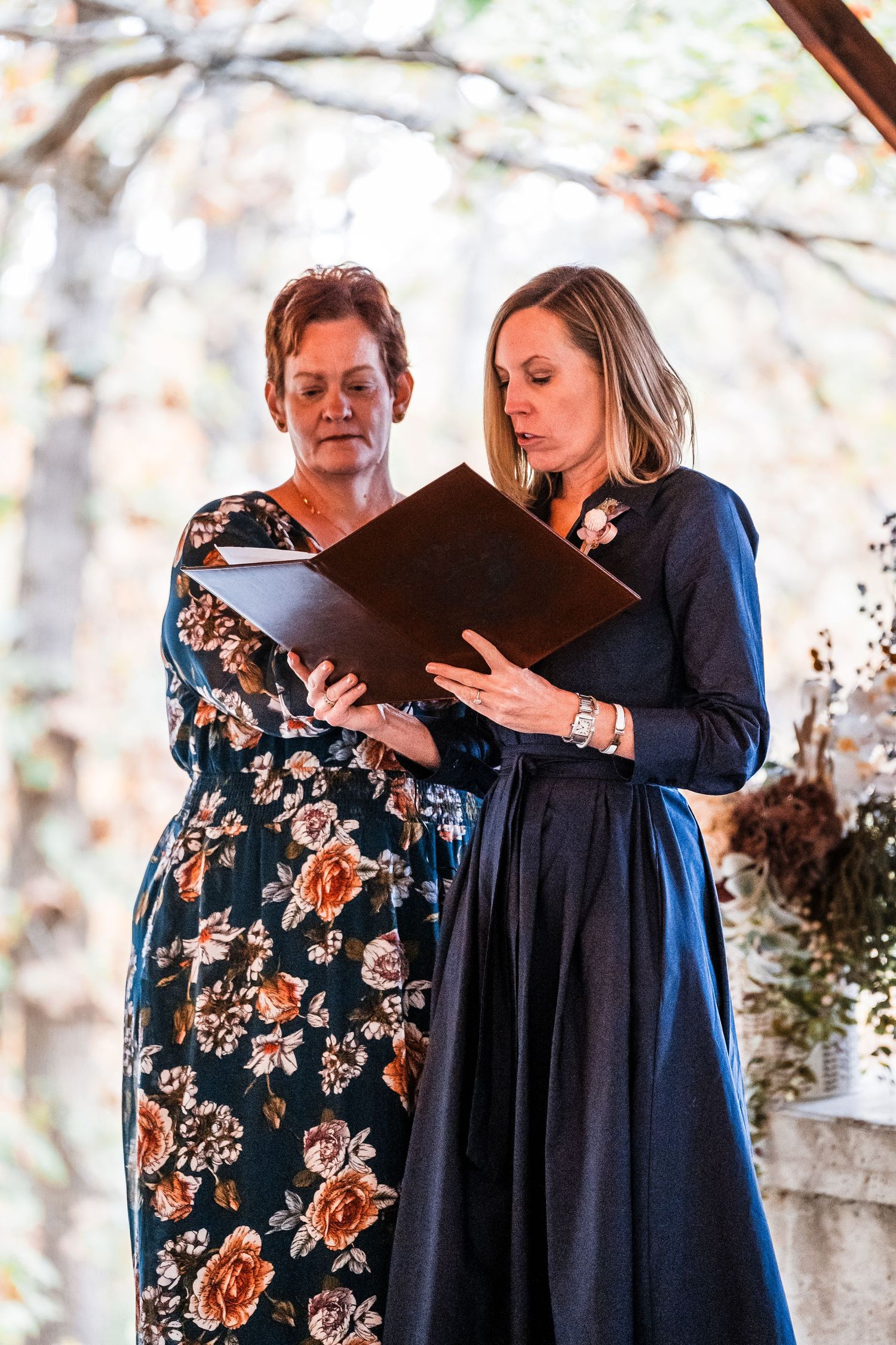 Two women in floral and navy dresses reading from folders during an outdoor ceremony.