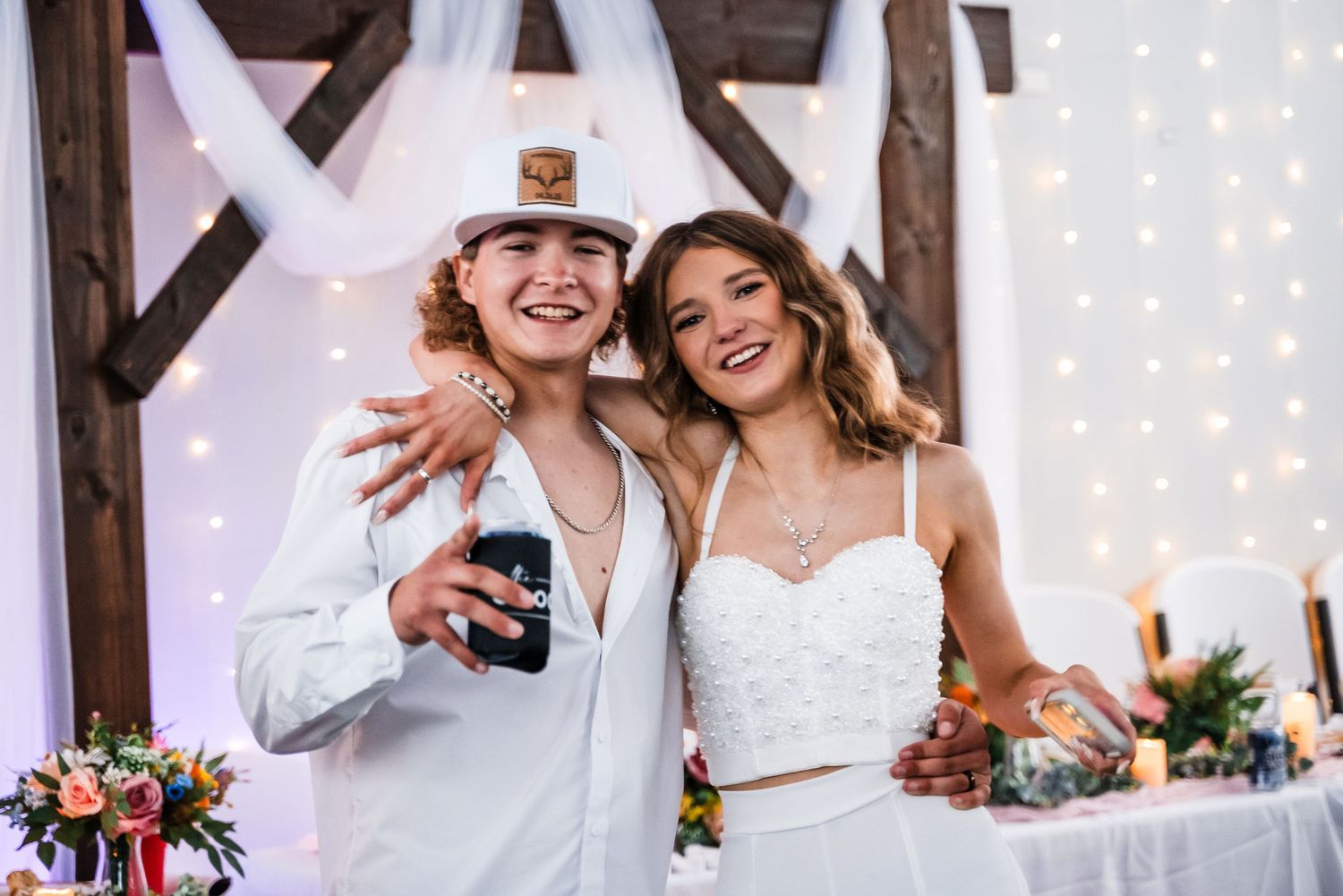 Two happy people celebrating at a wedding reception with twinkle lights and floral decorations in the background.