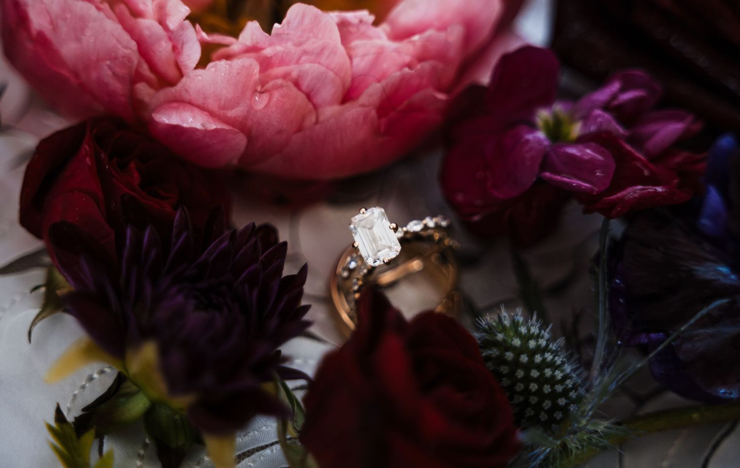 A diamond engagement ring nestled among colorful flowers including pink peonies and dark purple blooms.