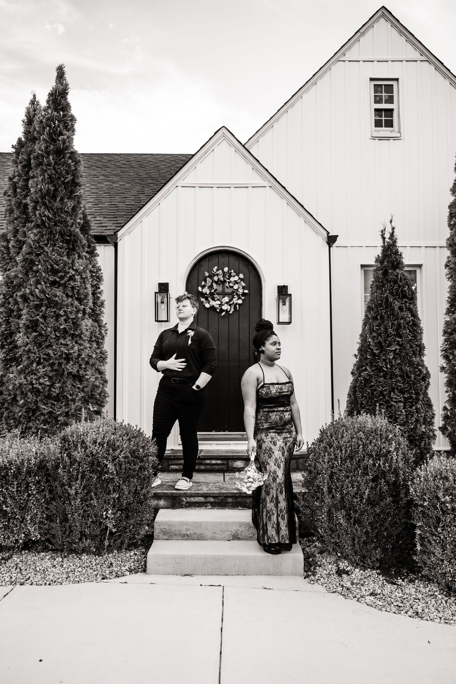A formal portrait of a couple standing on steps in front of a white church with tall cypress trees.