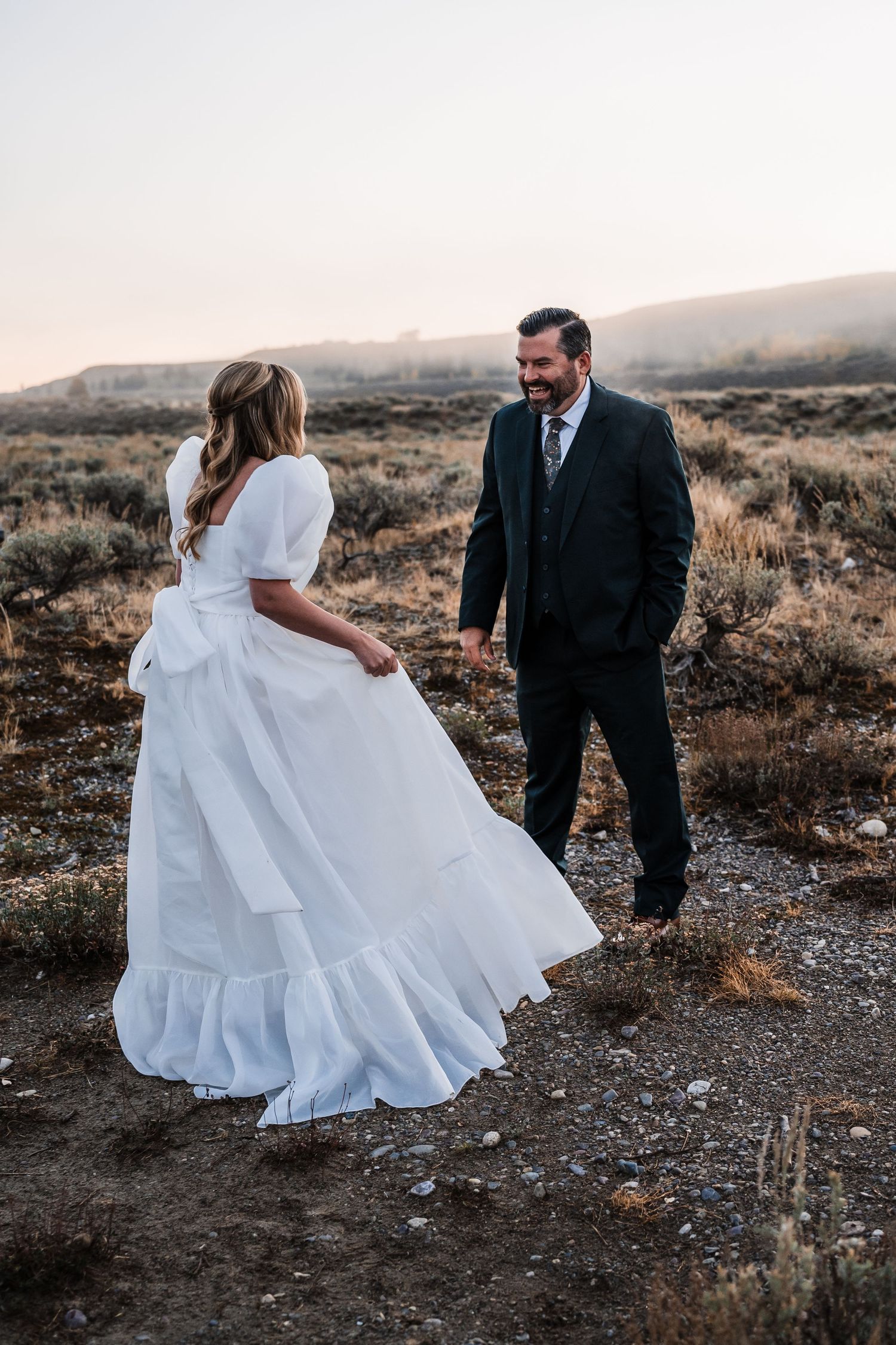 A couple takes romantic photos in a rustic outdoor setting with flowing white dress and dark suit.