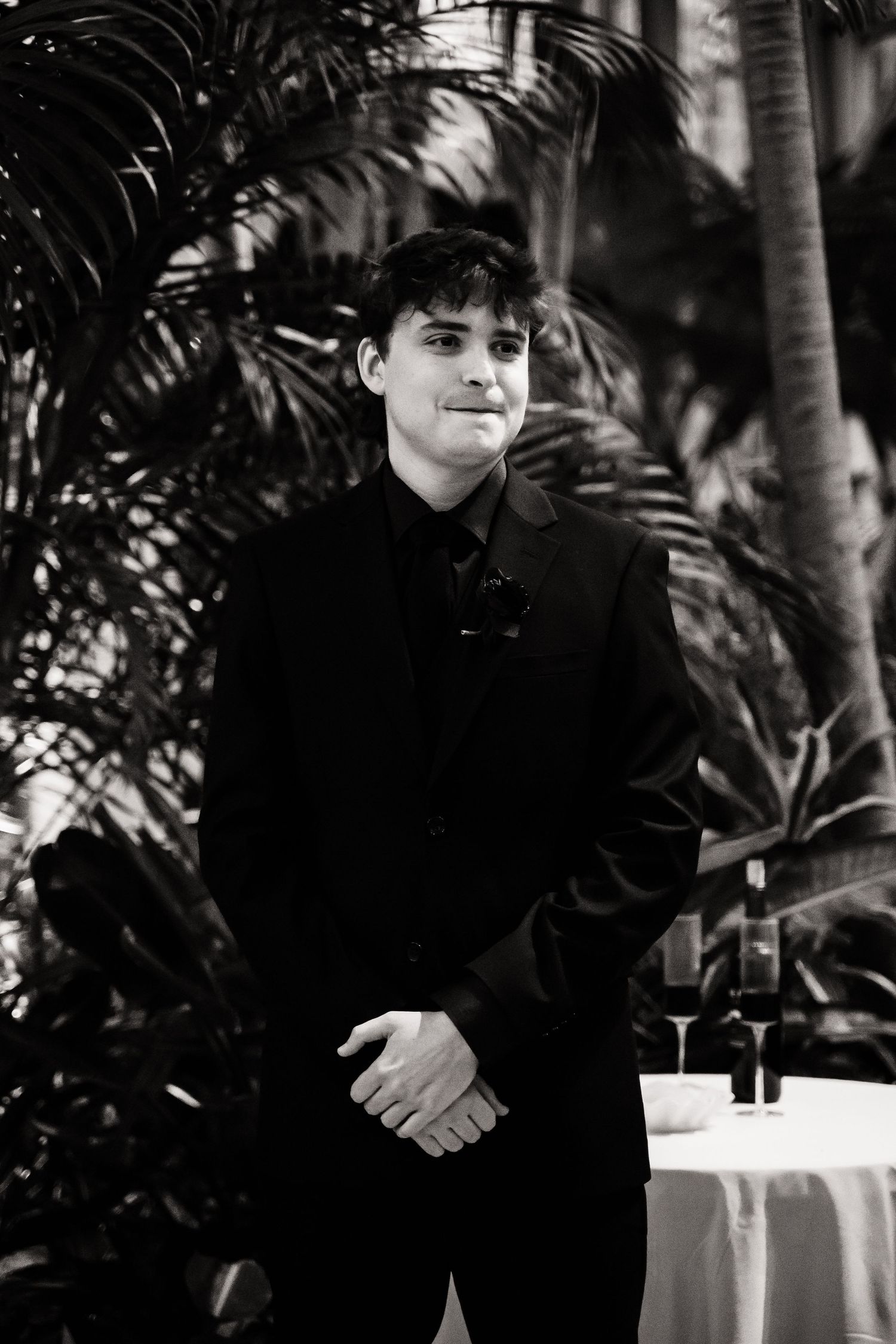 Black and white portrait of a person in formal attire standing near tropical plants.