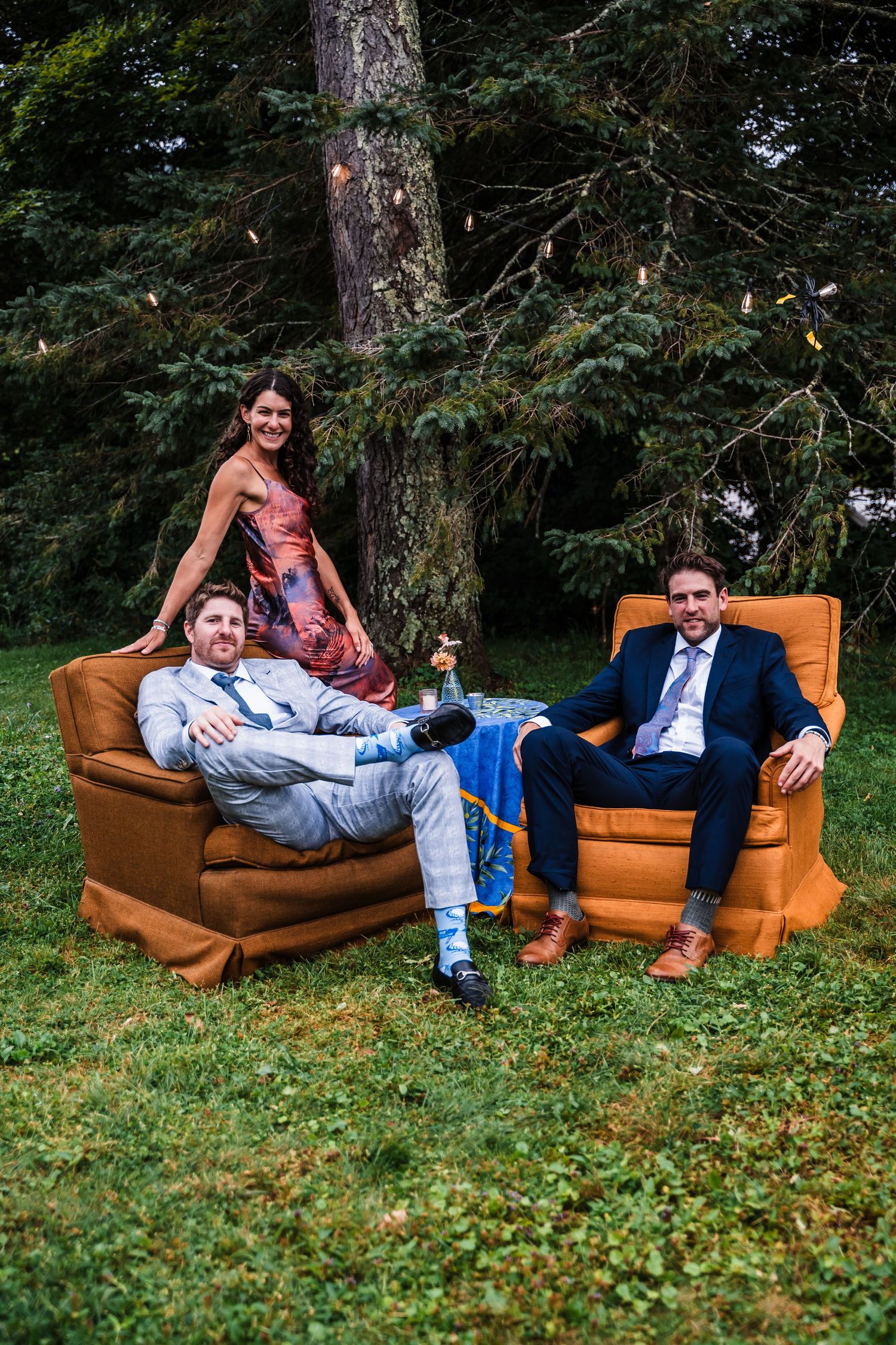 Wedding guests relaxing on vintage orange armchairs outdoors under tall trees.