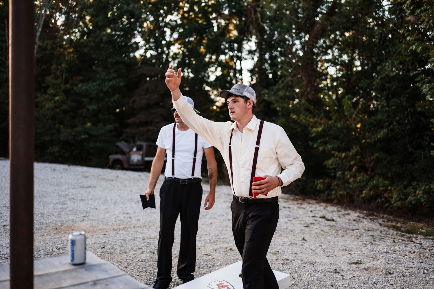 Two men in suspenders and dress shirts dance outdoors on a gravel path at dusk.