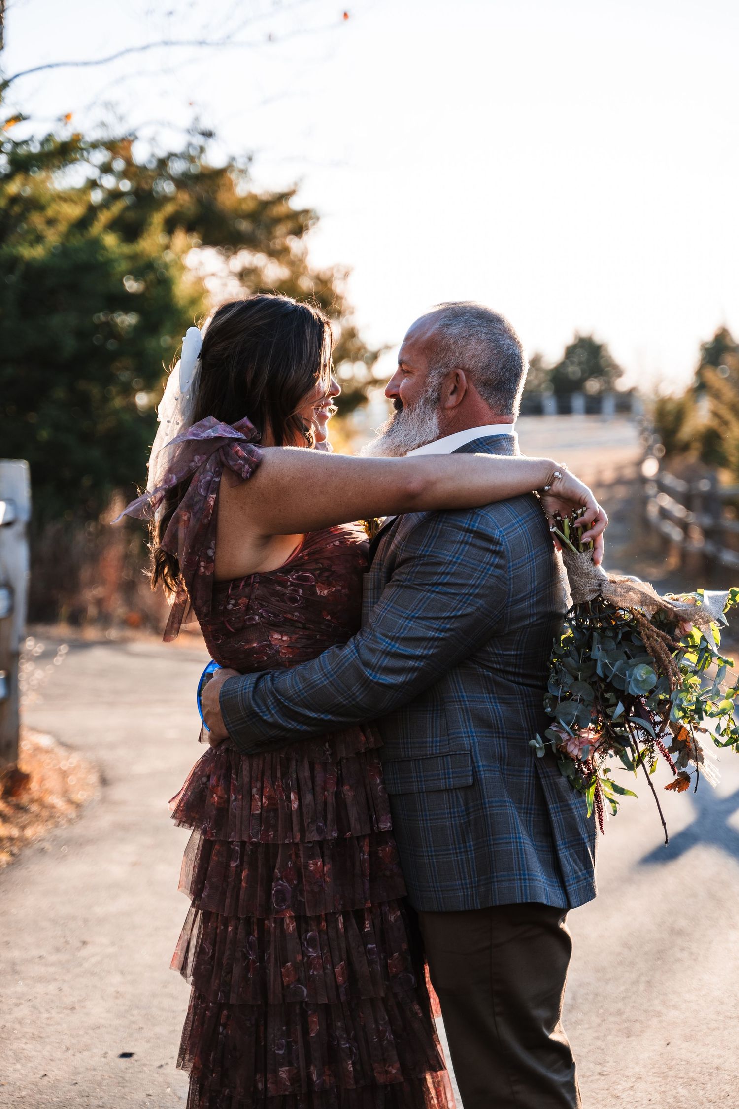 Couple in formal attire embracing during sunset by lake with floral bouquet.
