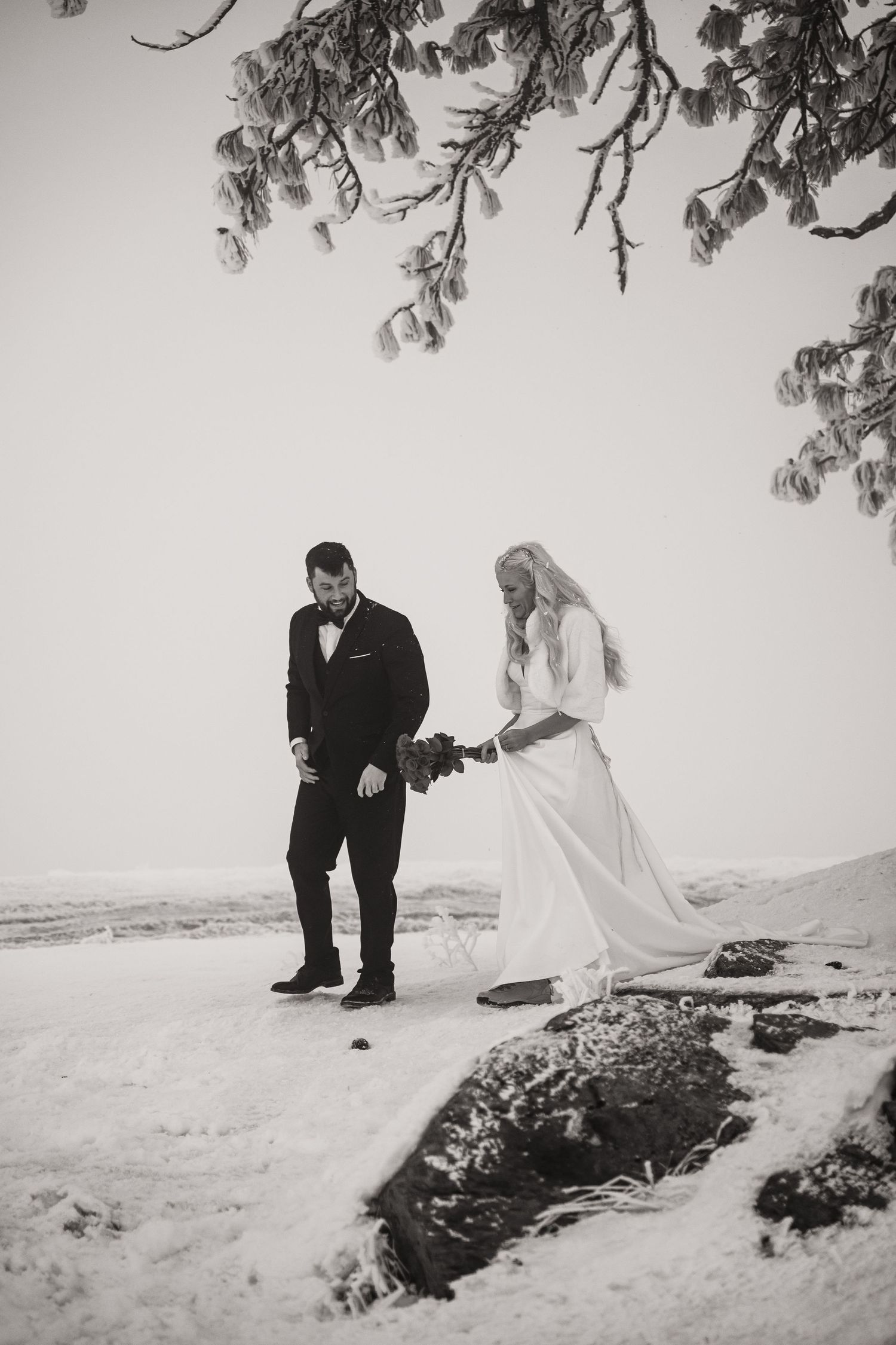 Black and white photo of wedding couple walking through snow under frosted branches.