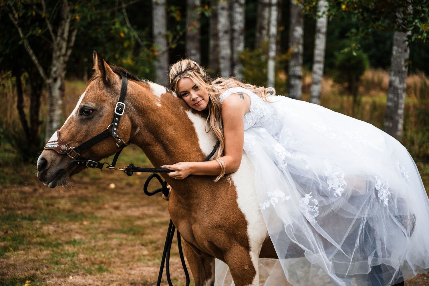A brown horse stands in a field while someone in a white wedding dress leans against it.
