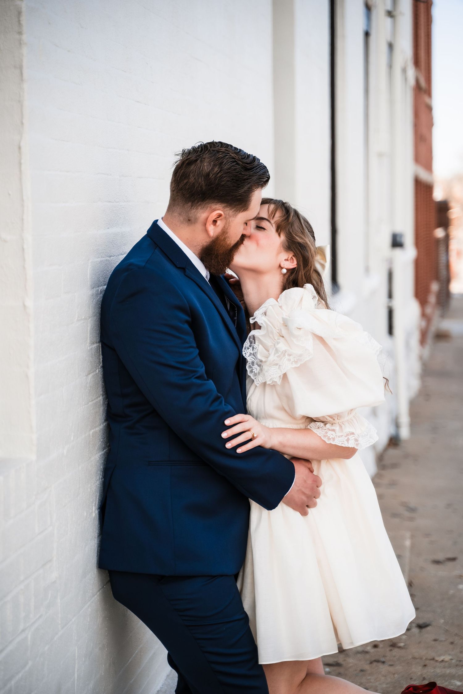 Romantic moment between couple against white wall in vintage-inspired wedding attire.