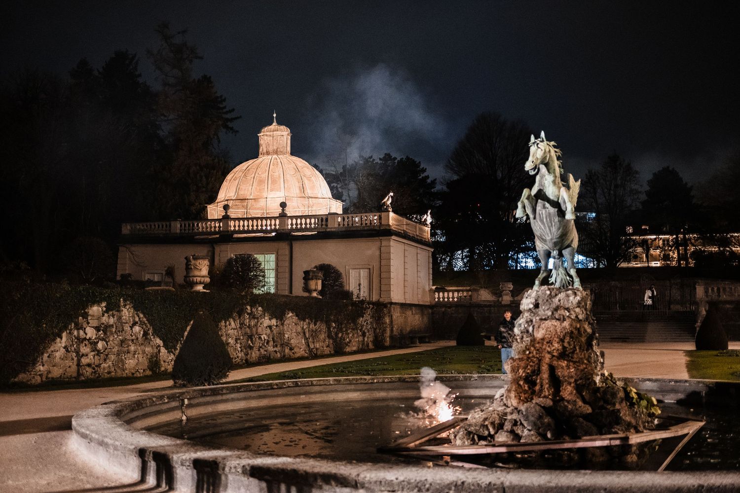 Illuminated stone statues and architectural features create a dramatic nighttime scene in a park or garden setting.