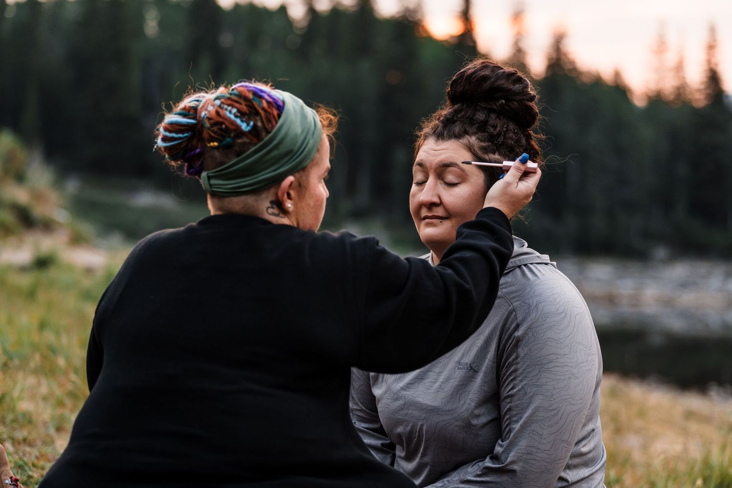 Two women sit outdoors near a forest lake at sunset, one applying makeup to the other's face.