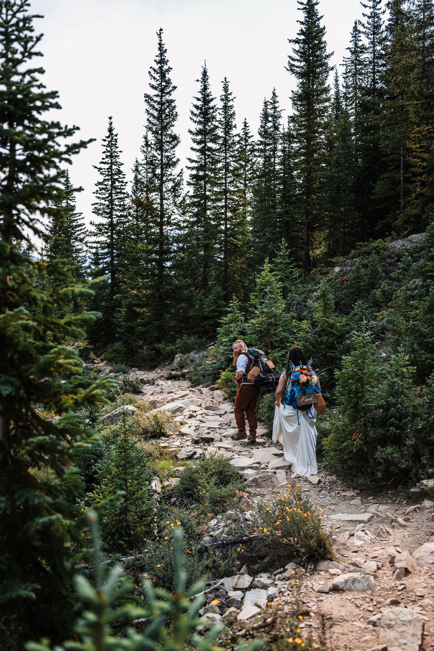 Bride in white dress and groom with backpacks hike rocky mountain trail through pine forest on elopement day.