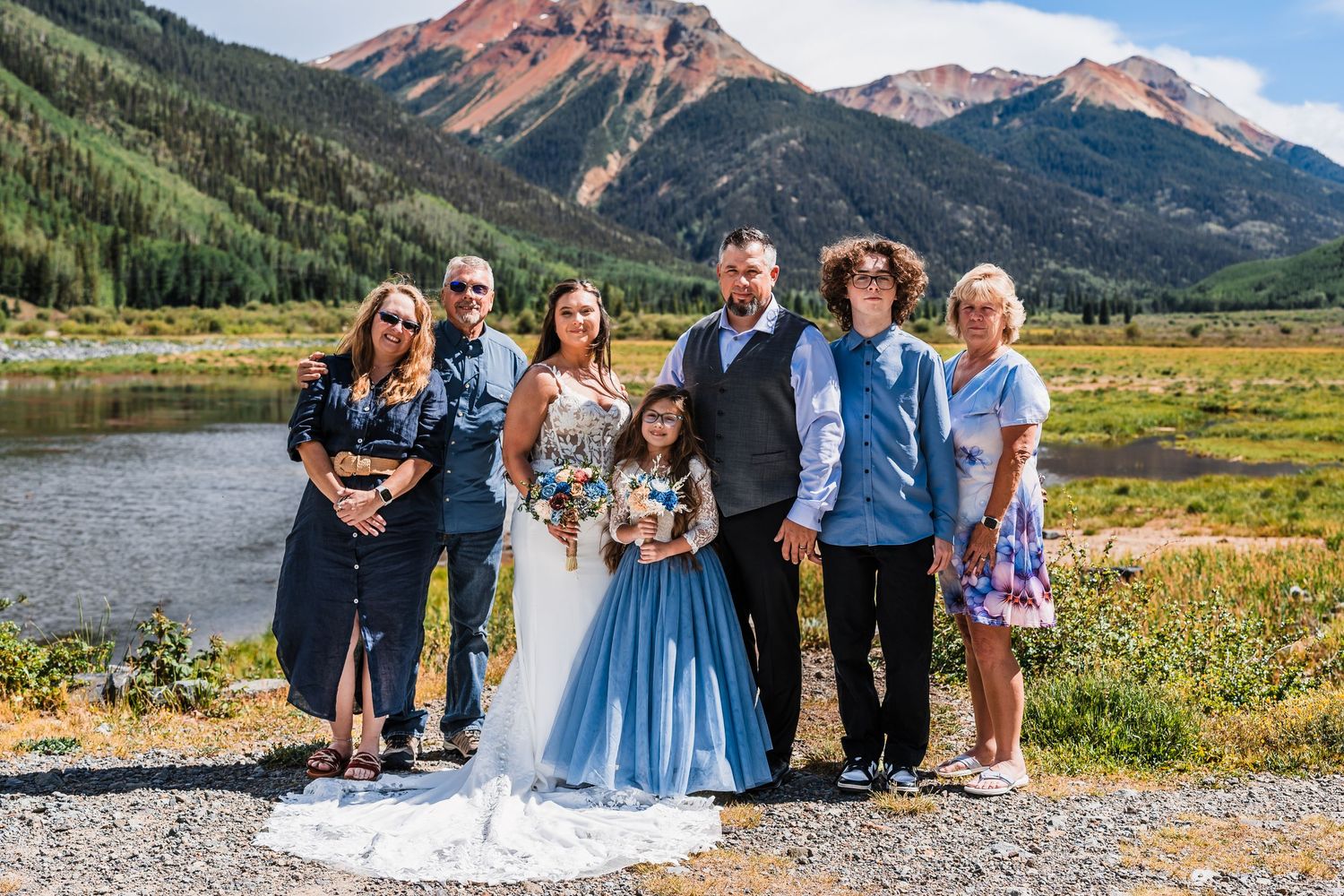Wedding party group photo with bride, groom, and family posing before a scenic mountain lake backdrop.