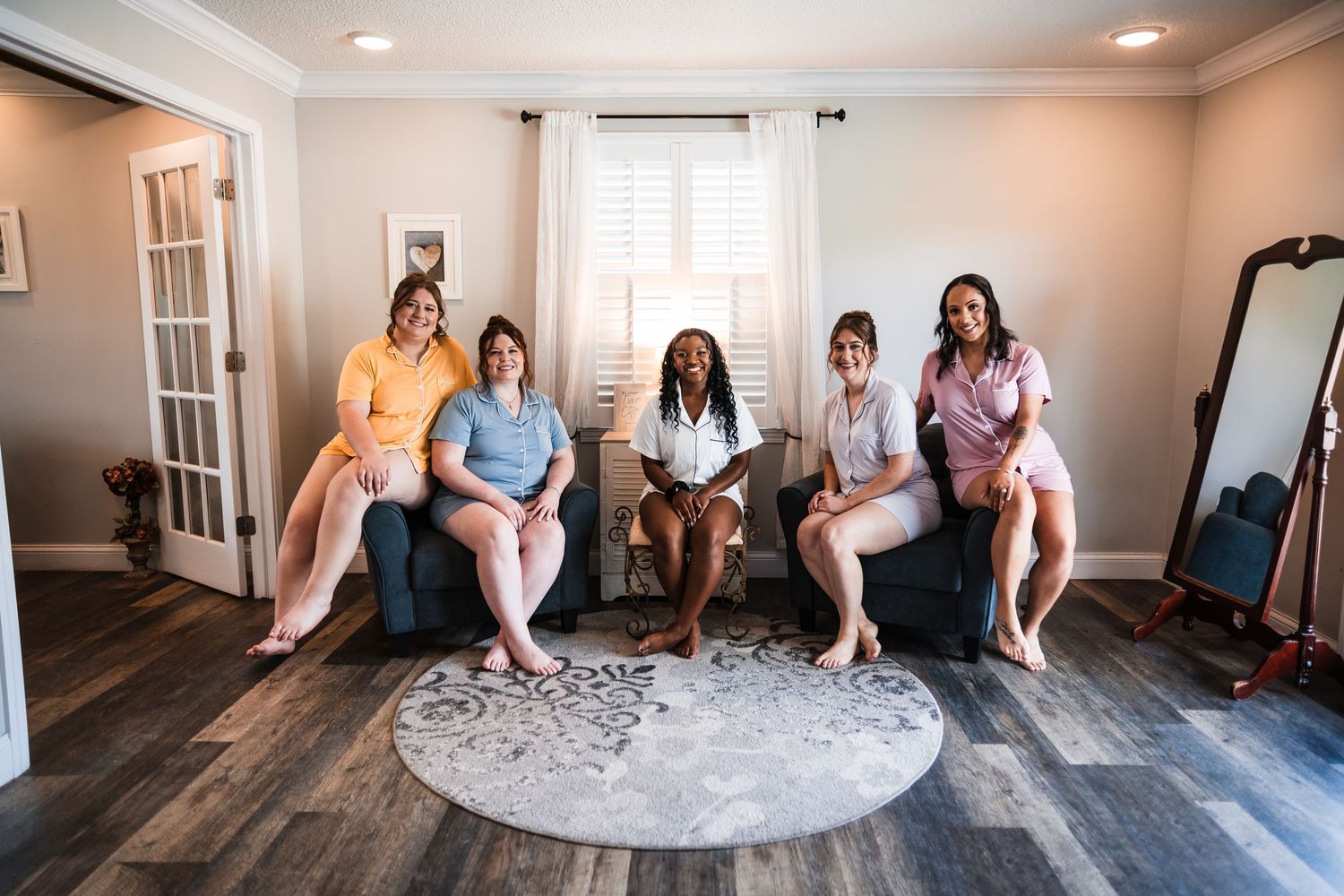 Five women in pastel pajamas smile together on chairs in a bright room during a bridal party getting ready session.