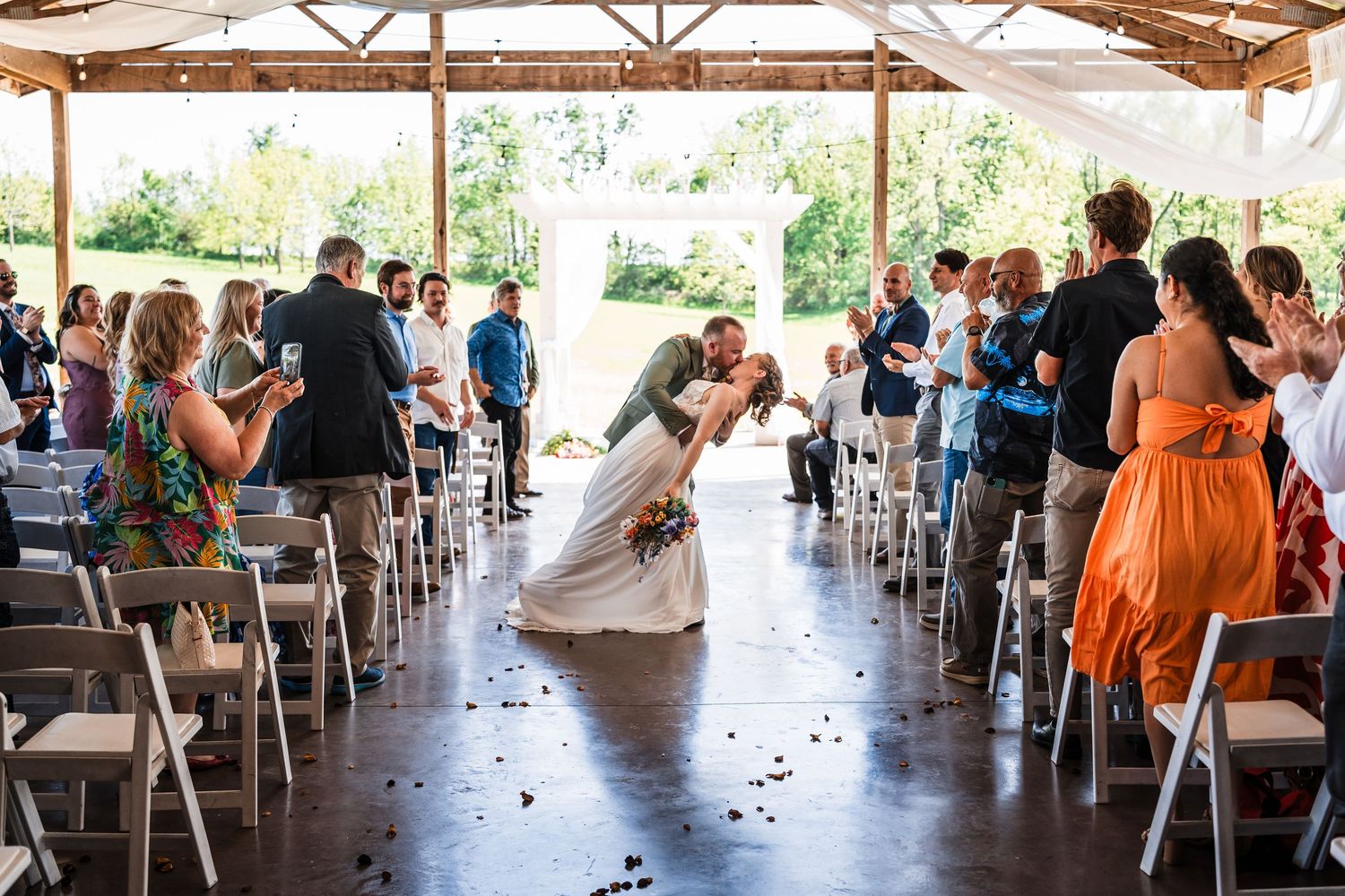 Bride and groom share romantic dip kiss while guests applaud in rustic outdoor ceremony.