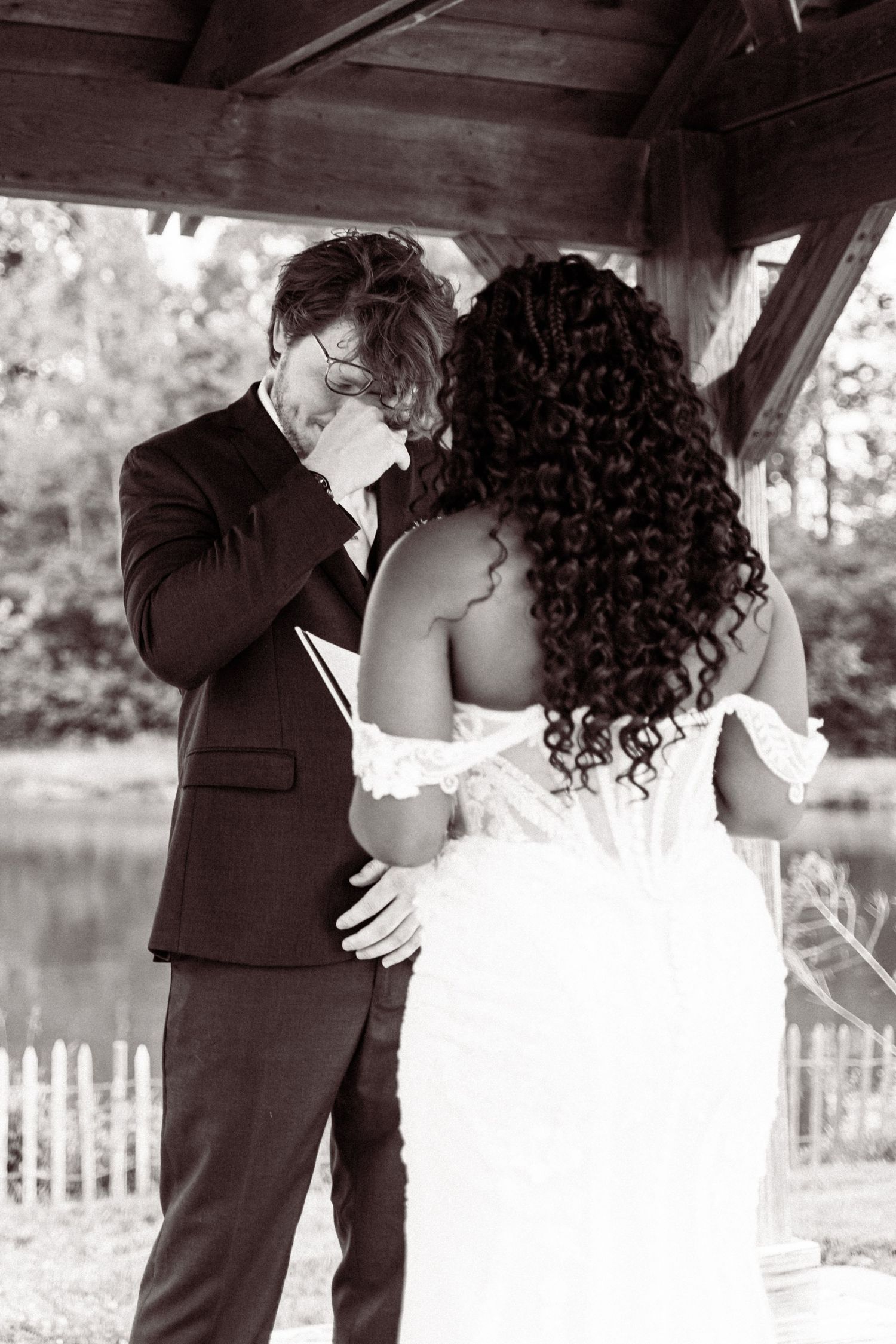 Emotional groom wipes tears during first look with bride in lace dress under rustic outdoor pavilion, black and white.