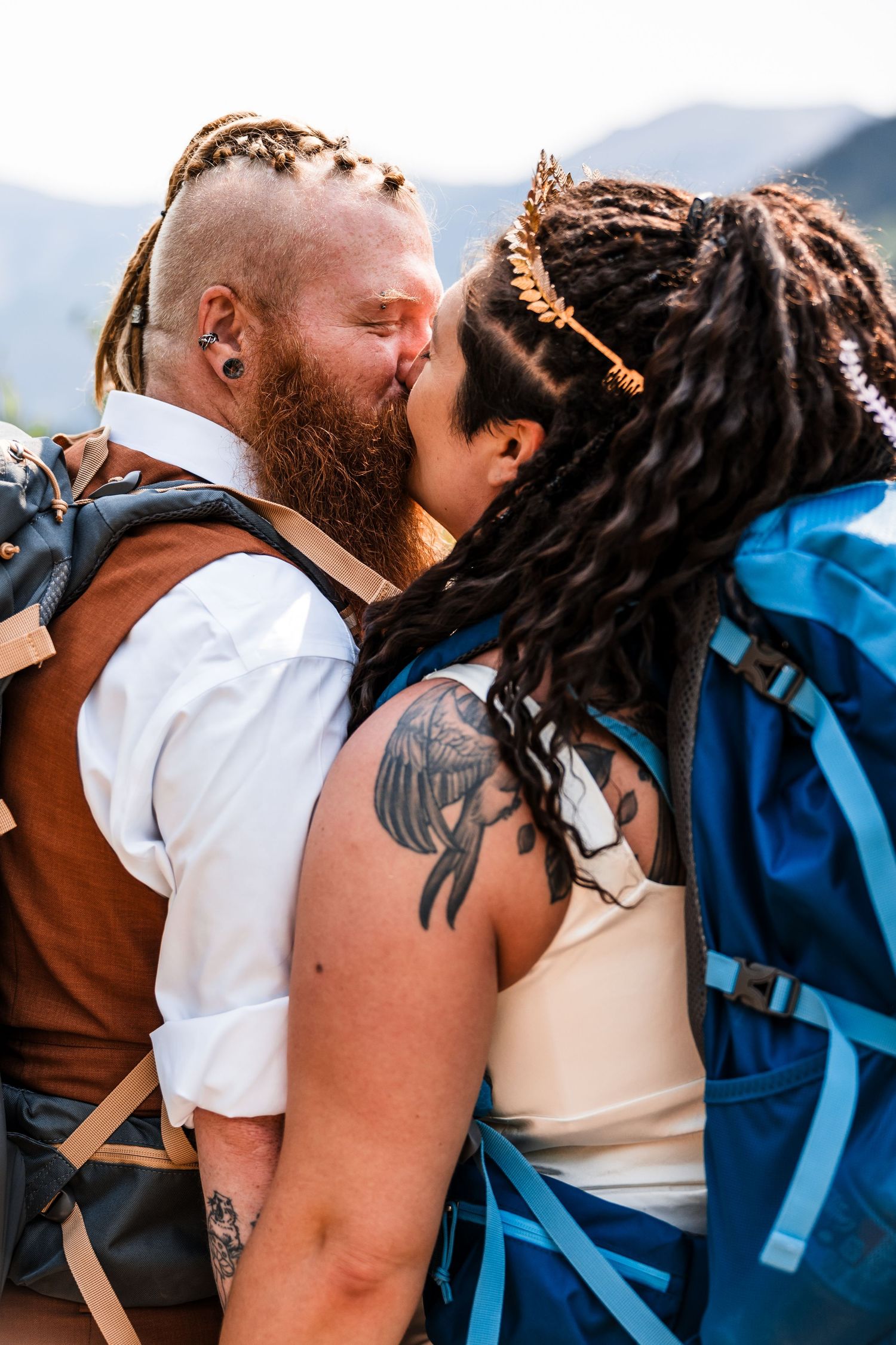 Tattooed couple shares first kiss with hiking backpacks on mountain elopement day.