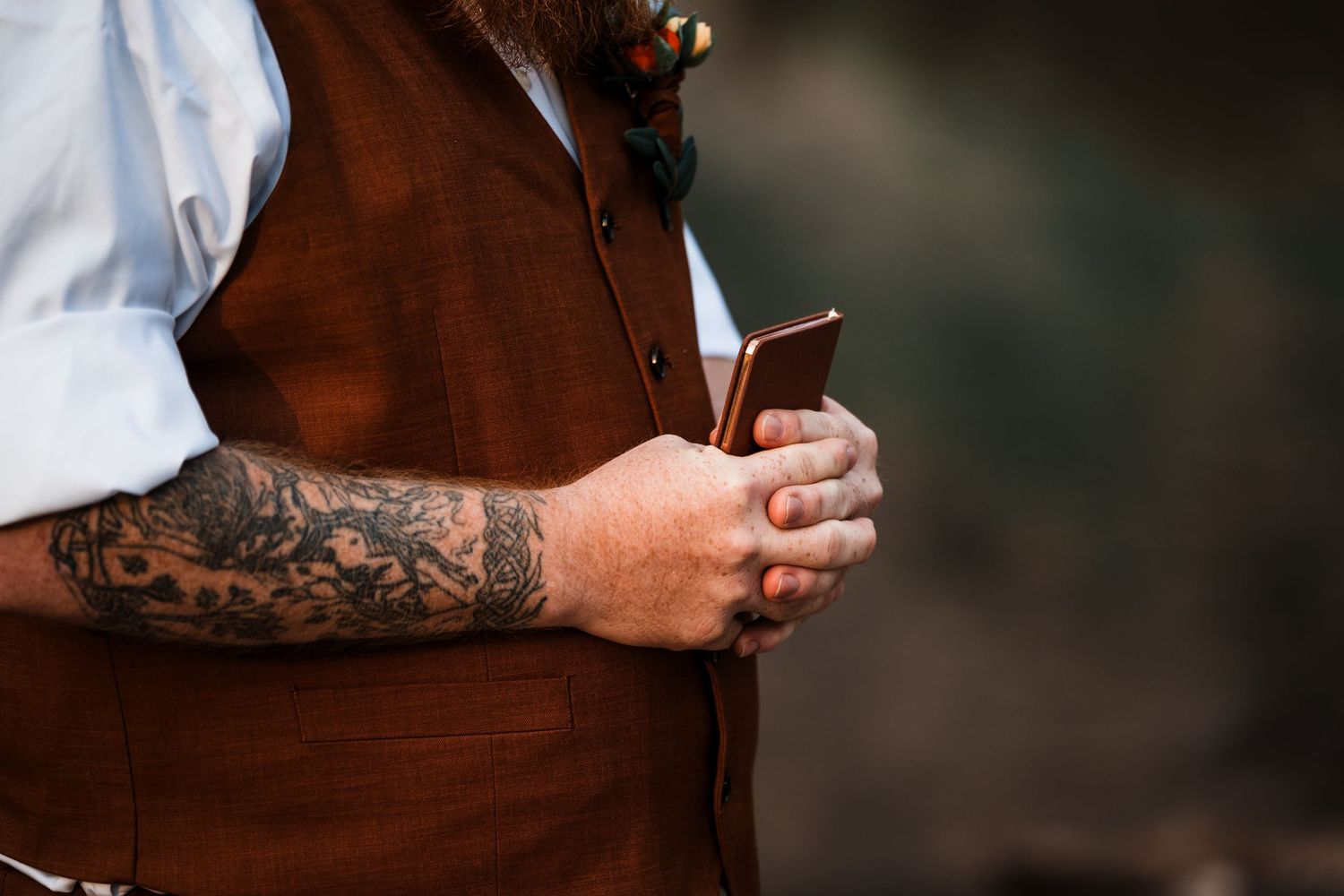 Close-up of tattooed groom in brown vest holding a small book during an outdoor wedding ceremony.