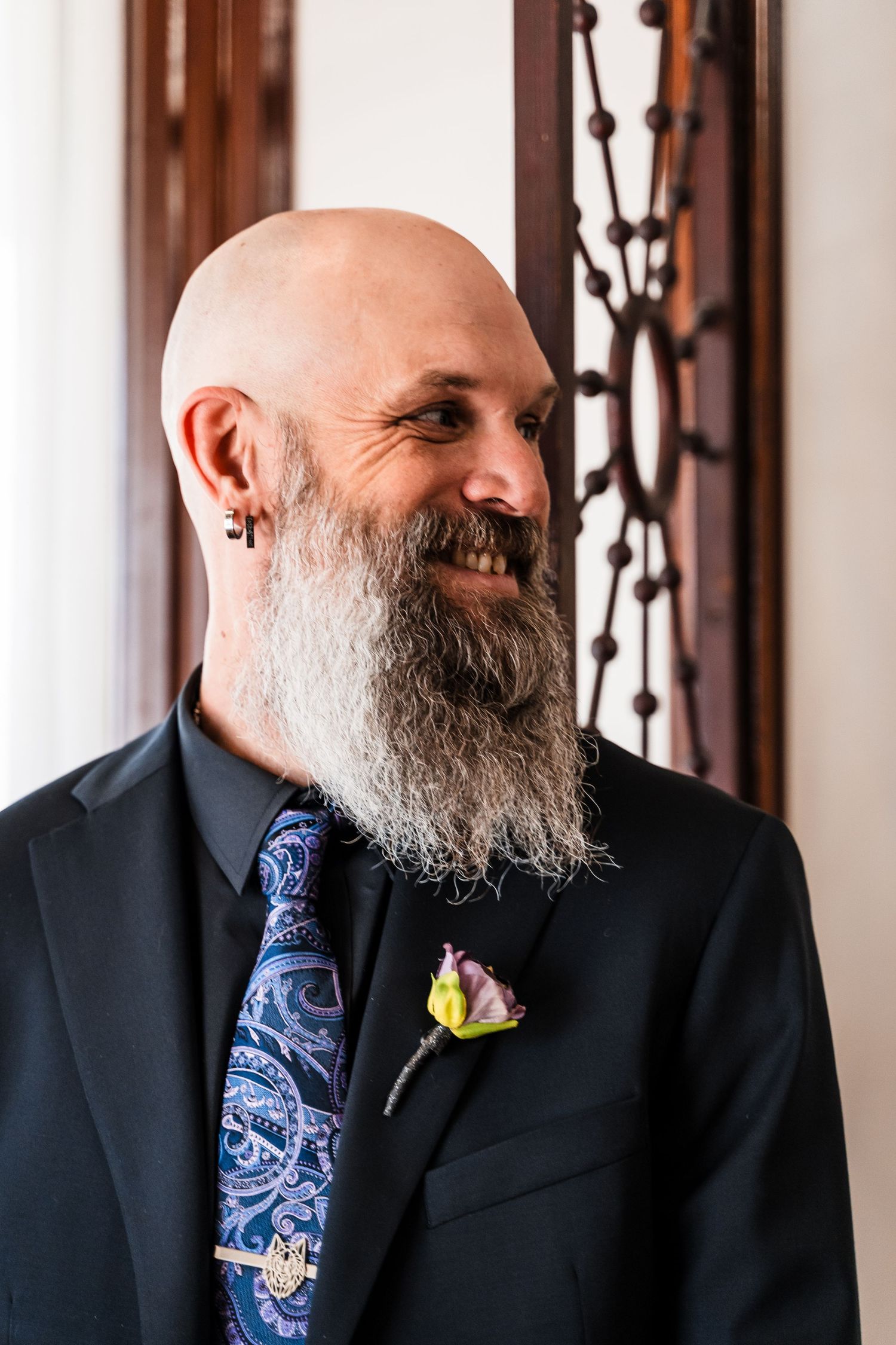Smiling bald groom with long gray beard wearing black suit, blue paisley tie, and floral boutonniere.