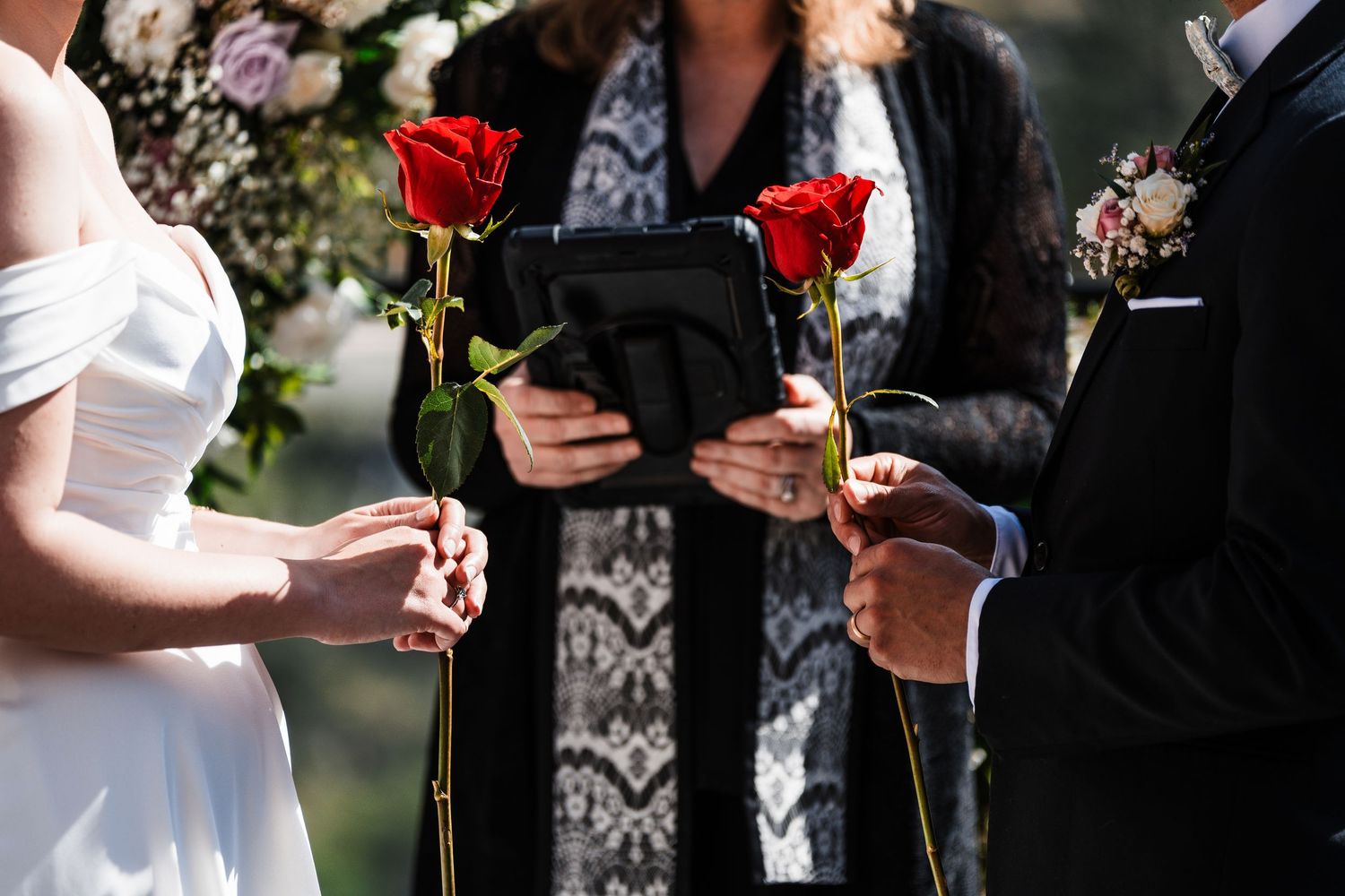 Bride and groom each holding red roses during wedding ceremony with officiant reading vows.