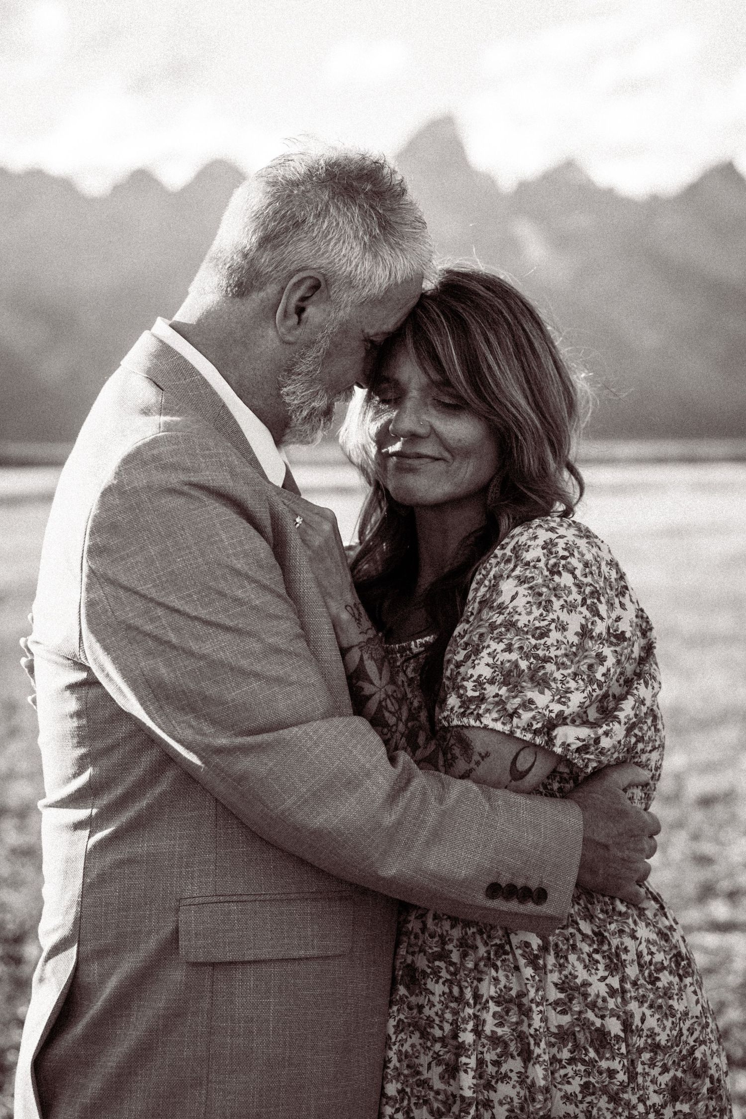 A romantic black and white portrait of a couple embracing outdoors with mountain peaks visible in the background.