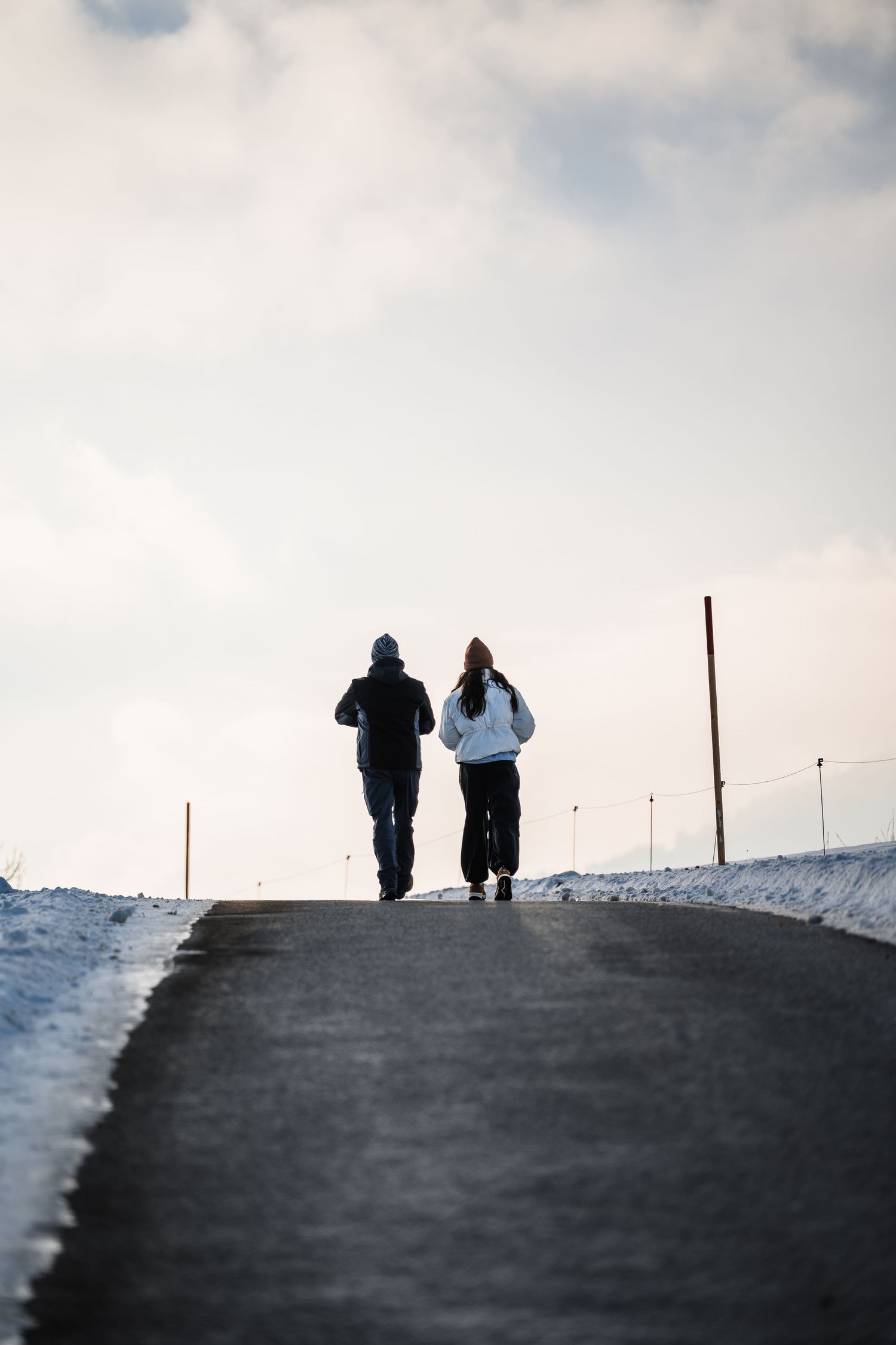 Two people walking together on a snowy road against a cloudy winter sky.