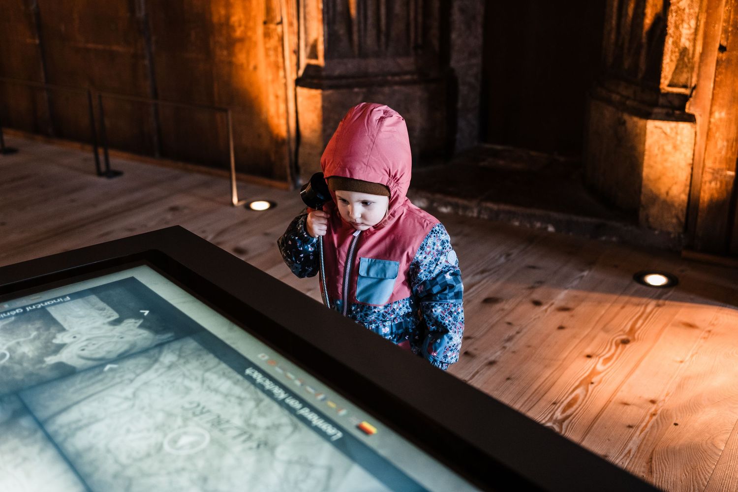 A child in a pink winter hat and blue patterned coat looks down at an ice fishing hole in a wooden floor.