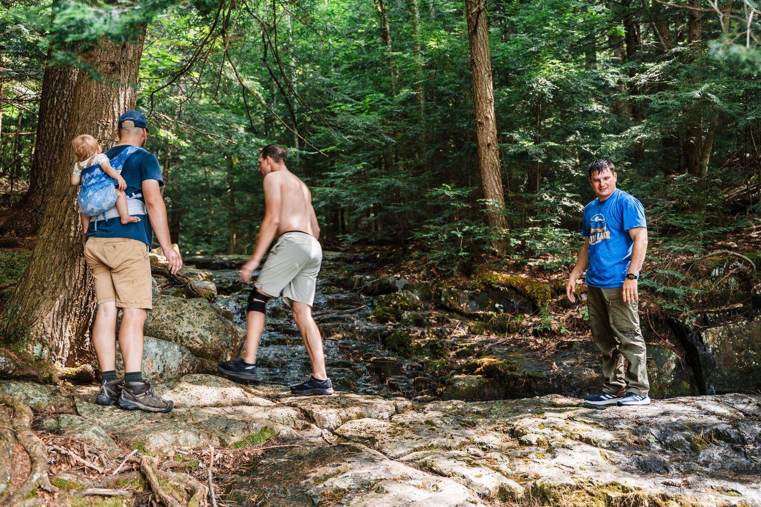 Three people explore a rocky stream bed surrounded by lush forest on a sunny summer day.