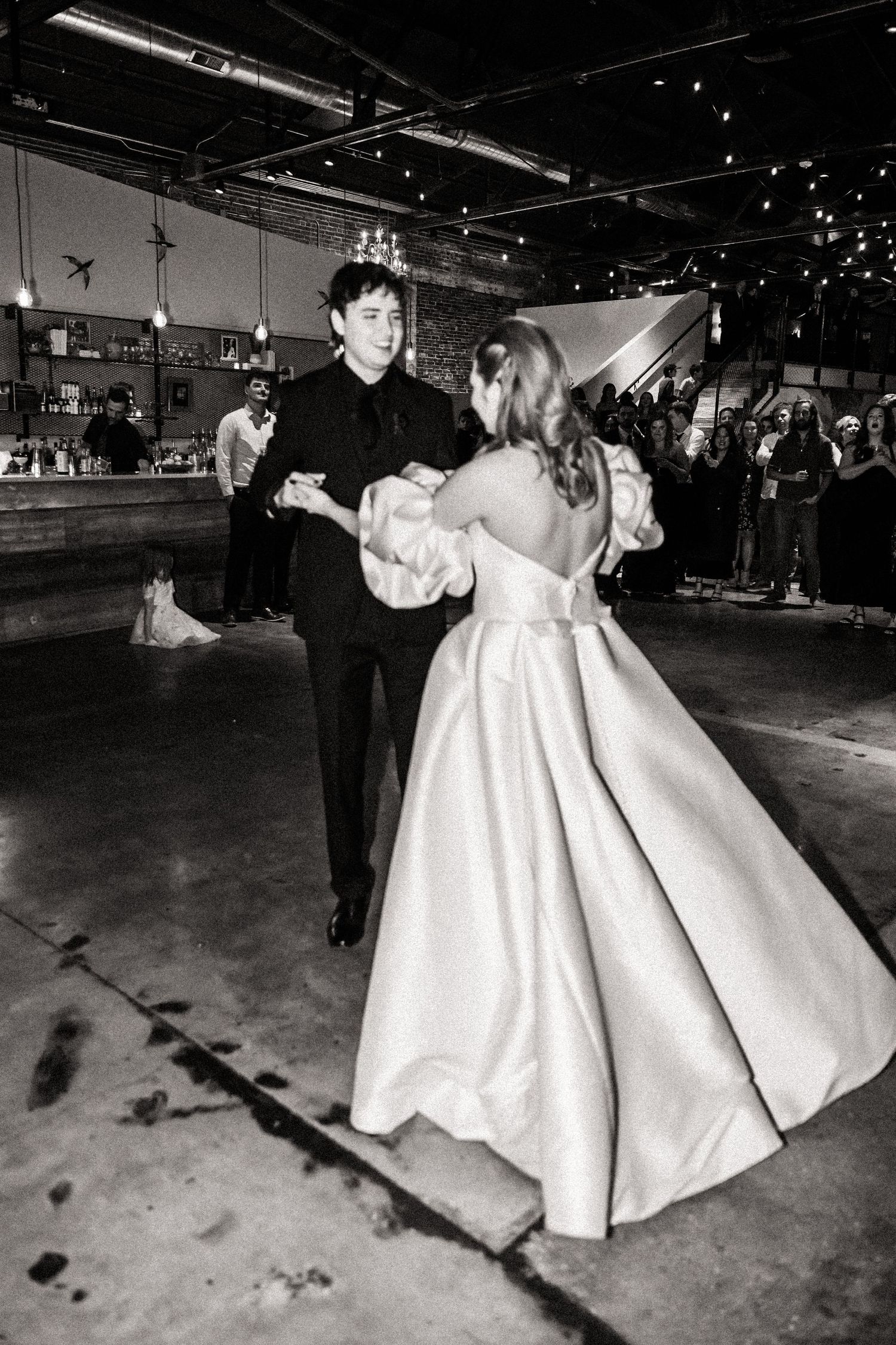A couple dances in a spacious industrial venue with exposed ceiling and string lights.