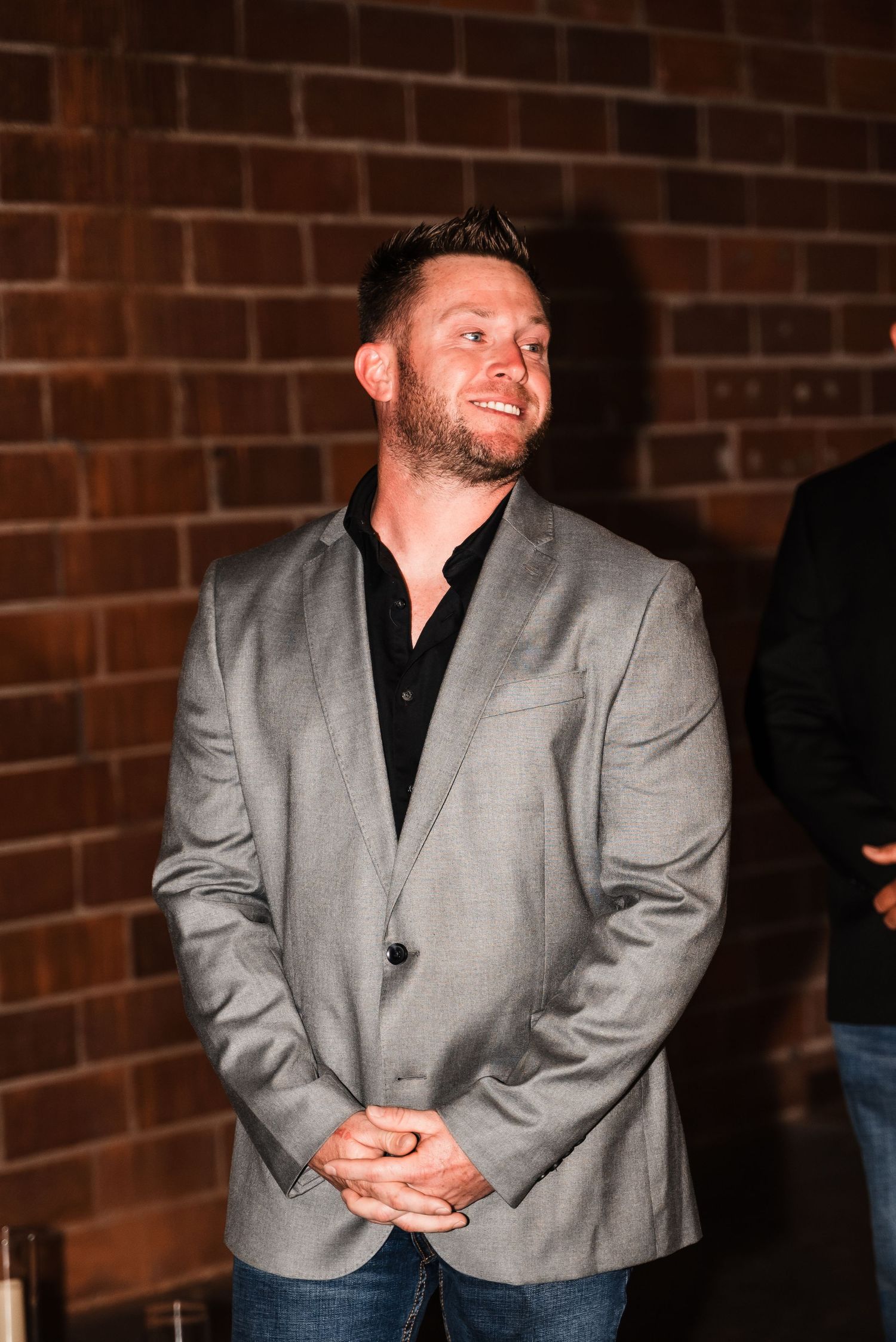 A person in a gray blazer and black shirt poses against a brick wall at an evening event.