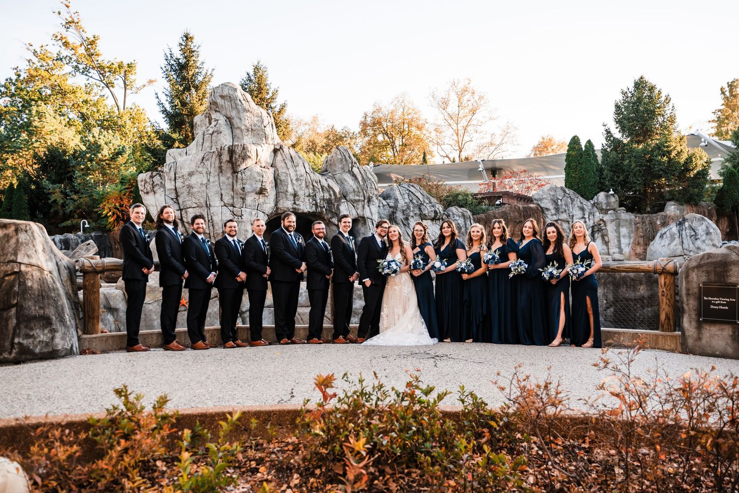 Wedding party in formal attire poses together against stone architecture during autumn.