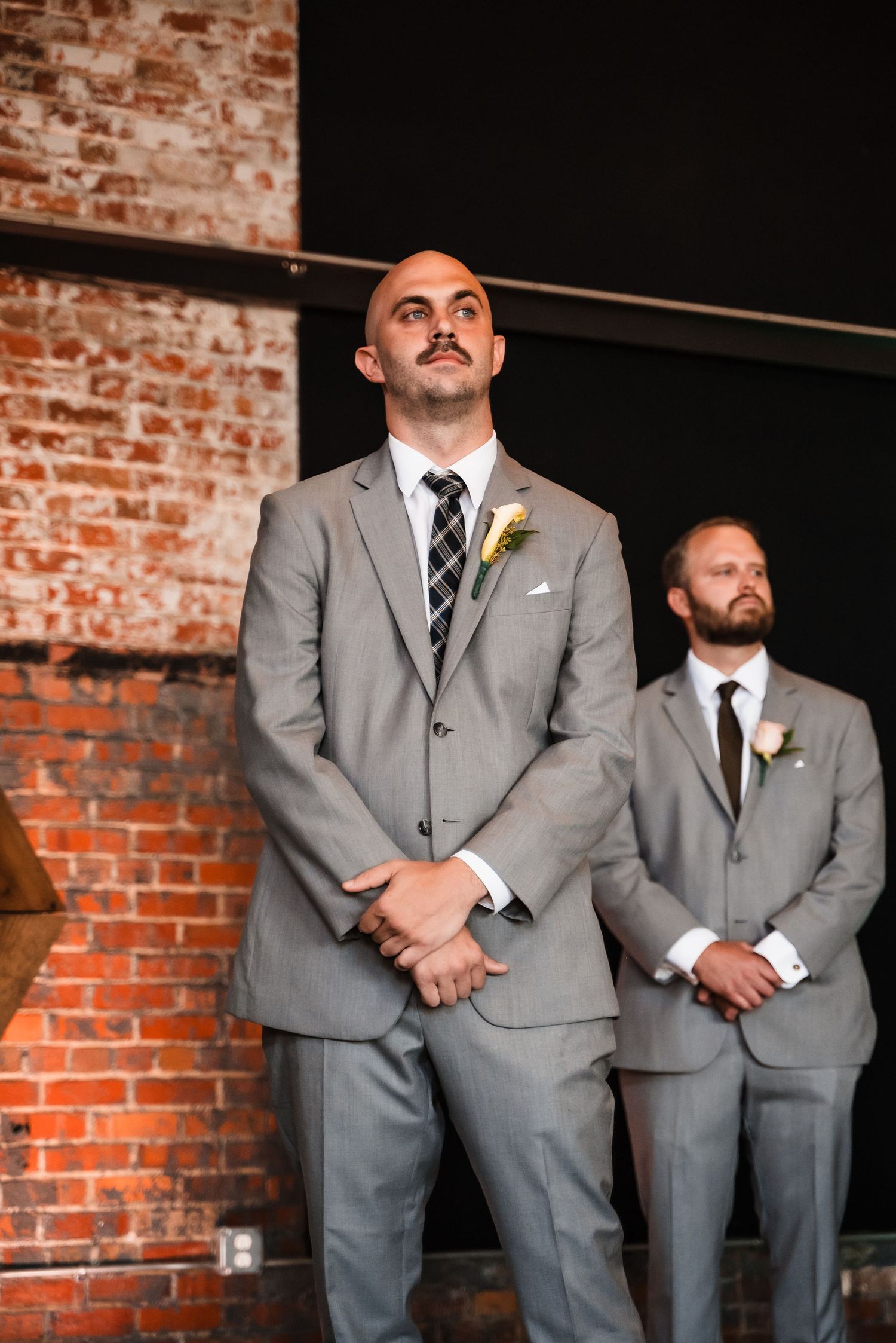 Two groomsmen in grey suits stand at attention during a wedding ceremony against exposed brick walls.