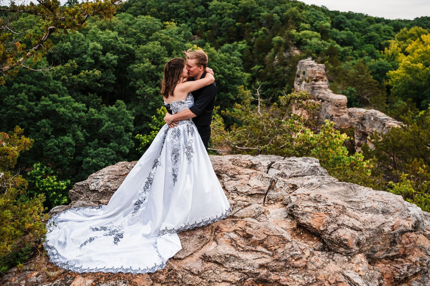 Wedding couple embracing on rocky cliff with dramatic forest backdrop and flowing white dress.