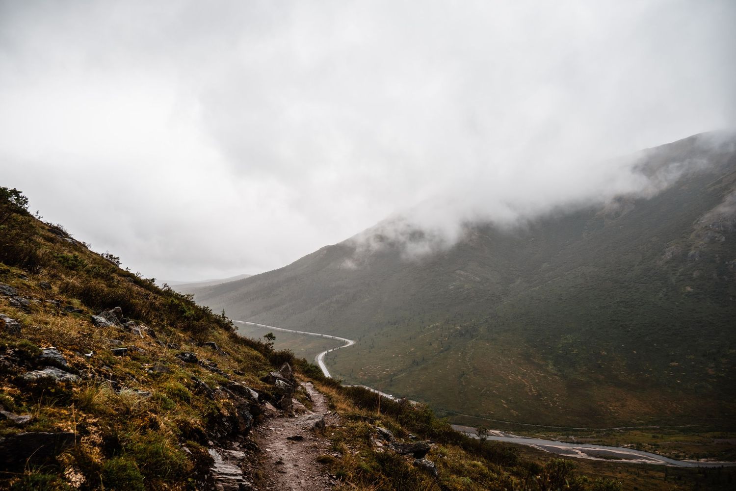 A misty mountain hiking trail winds through rugged Scottish highlands with a winding river valley below on a cloudy day.