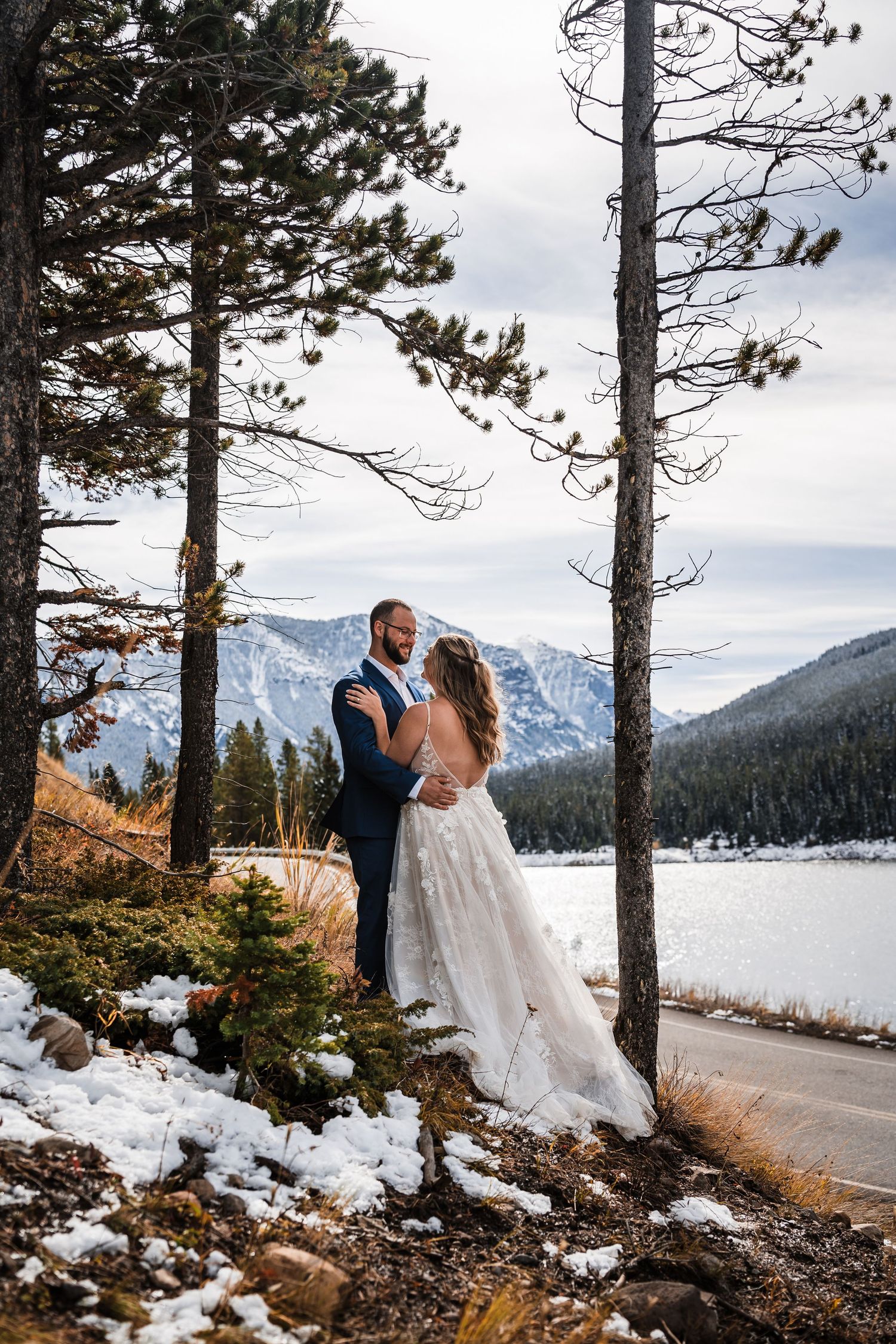 A romantic wedding portrait against a snowy mountain landscape framed by tall pine trees.